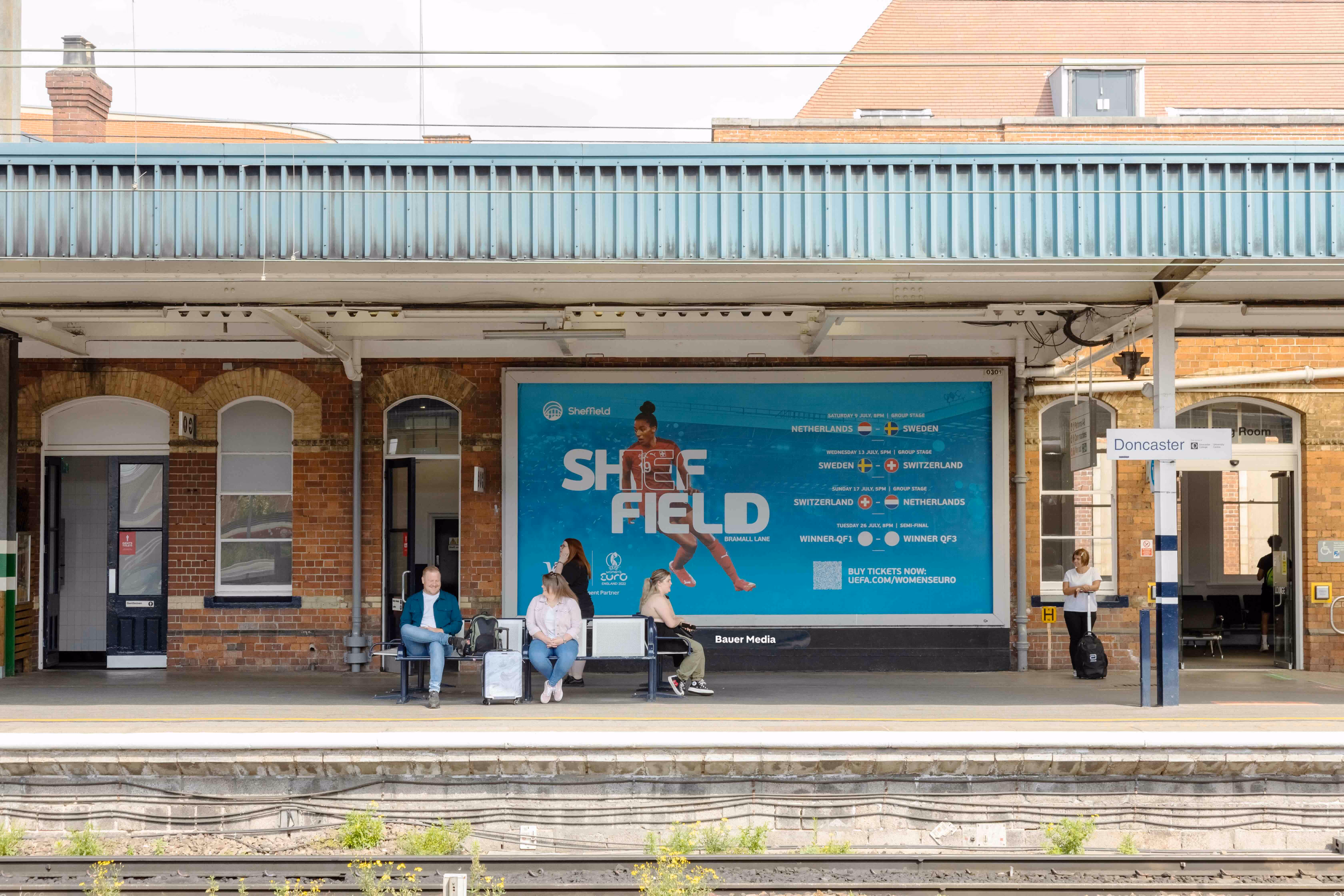 Bauer Media Outdoor billboard on the platform at Doncaster Station displays a blue Sheffield tourism ad featuring an athlete, as passengers wait for their train.