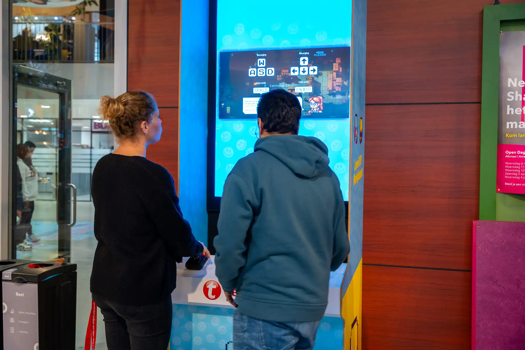Twee mensen staan bij een interactieve arcade-achtige spelconsole met een blauw verlichte scherm dat instructies toont voor bewegingen met de toetsen WASD en pijltjestoetsen.