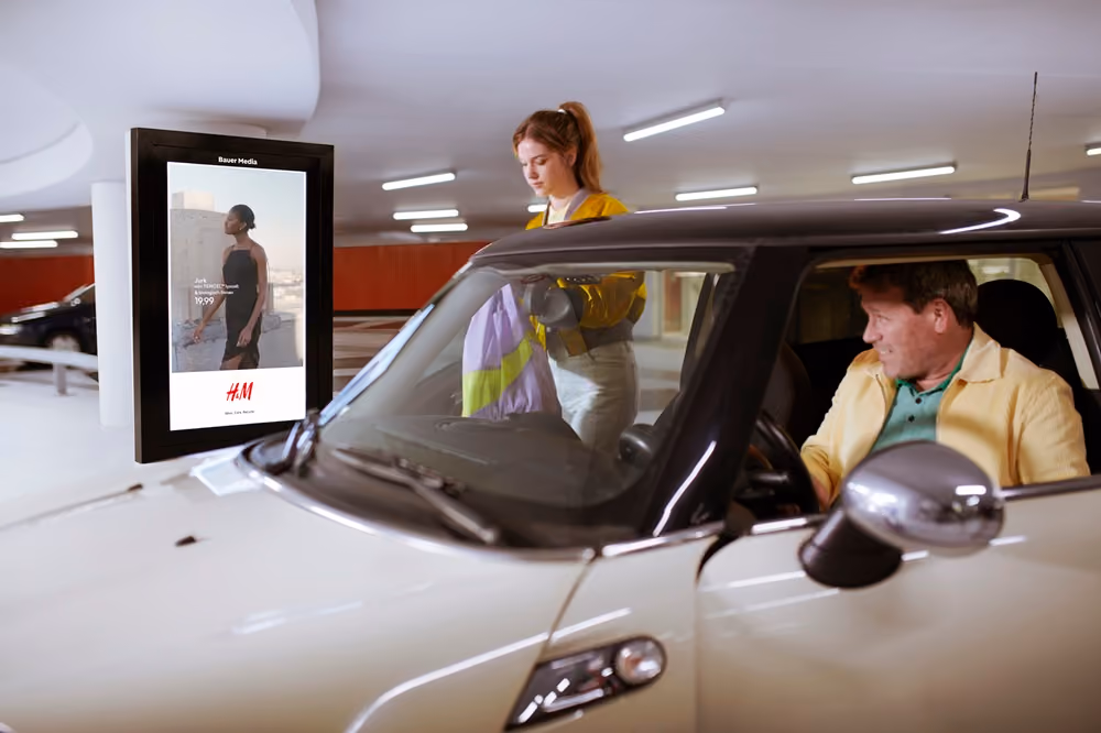 Man zit in een witte auto in een parkeergarage terwijl een vrouw naast de auto staat met een jas in haar handen.
