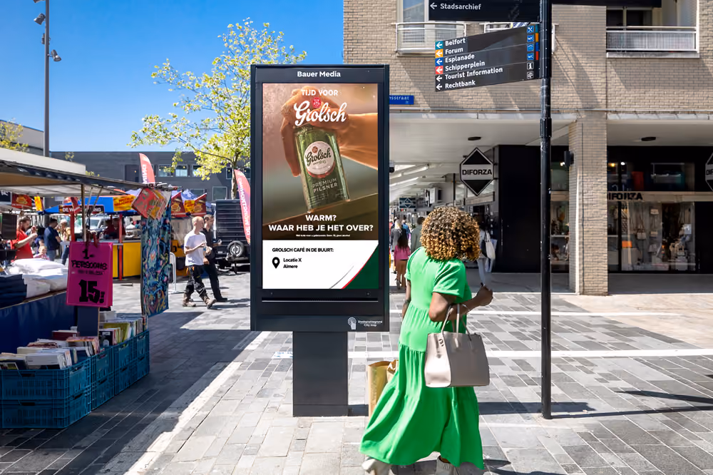 Vrouw in groene jurk loopt langs digitale reclame voor Grolsch bier op druk plein met winkels en marktkramen.