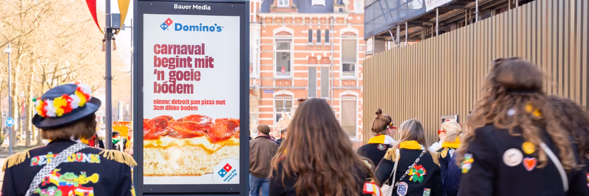 Street scene with people in colorful carnival costumes walking past a Domino's pizza ad in Dutch.
