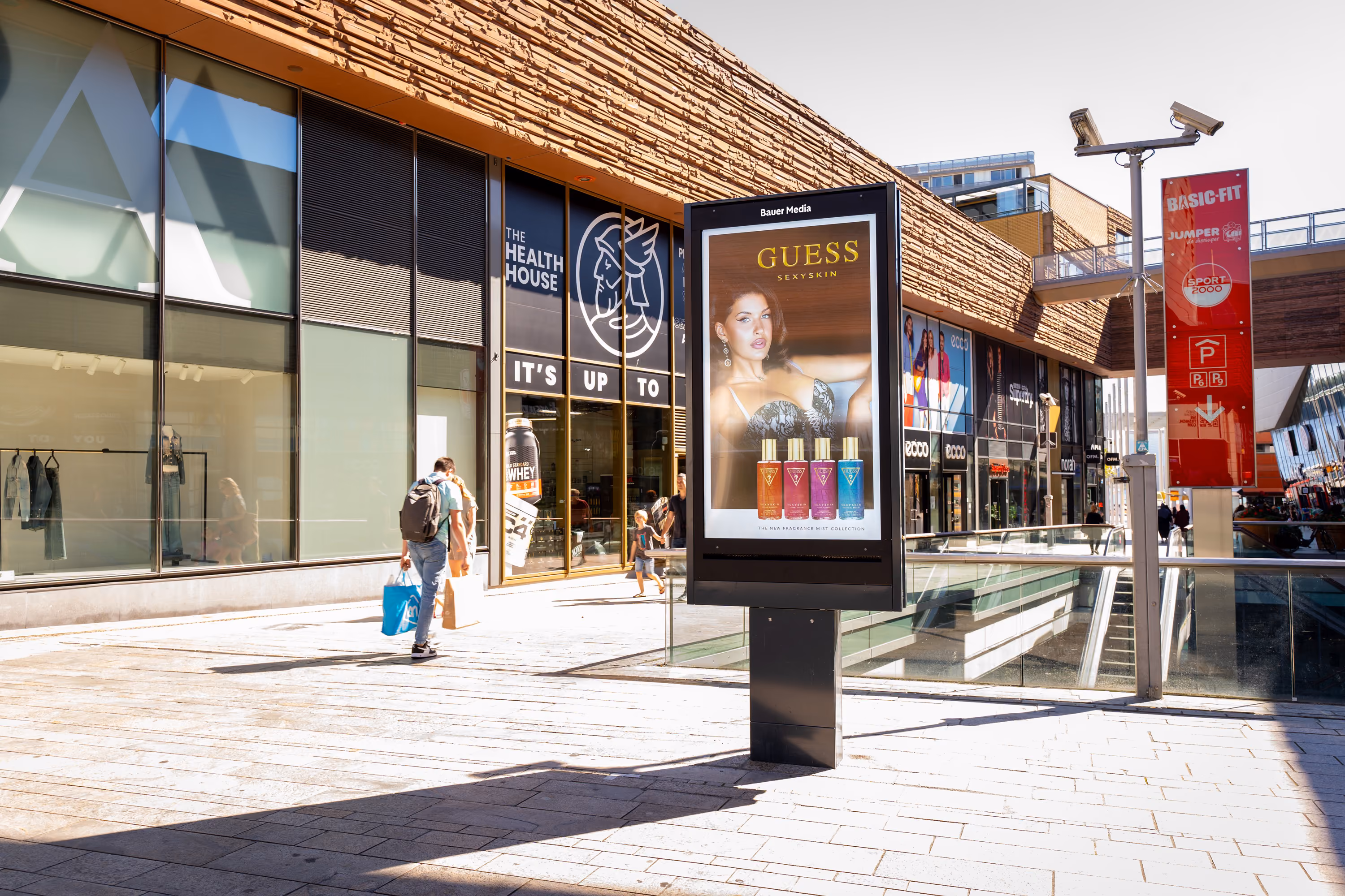 Straatbeeld van een winkelcentrum met een digitale reclamezuil die een Guess parfumadvertentie toont.