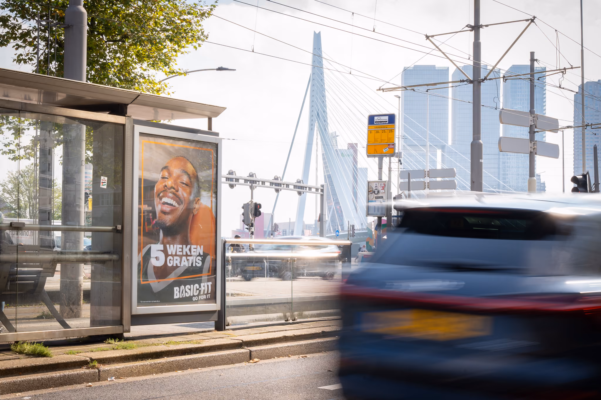 Bushalte met een Basic-Fit advertentie voor 5 weken gratis en de Erasmusbrug in Rotterdam op de achtergrond.