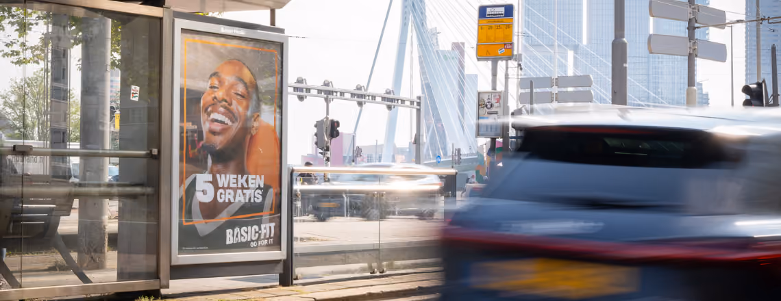 Reclamebord bij een bushalte met een glimlachende man en tekst '5 weken gratis Basic-Fit'.