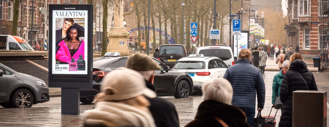 Drukke straat met mensen die lopen en auto's, met een Valentino-reclamebord en een grachtenpand in de achtergrond.