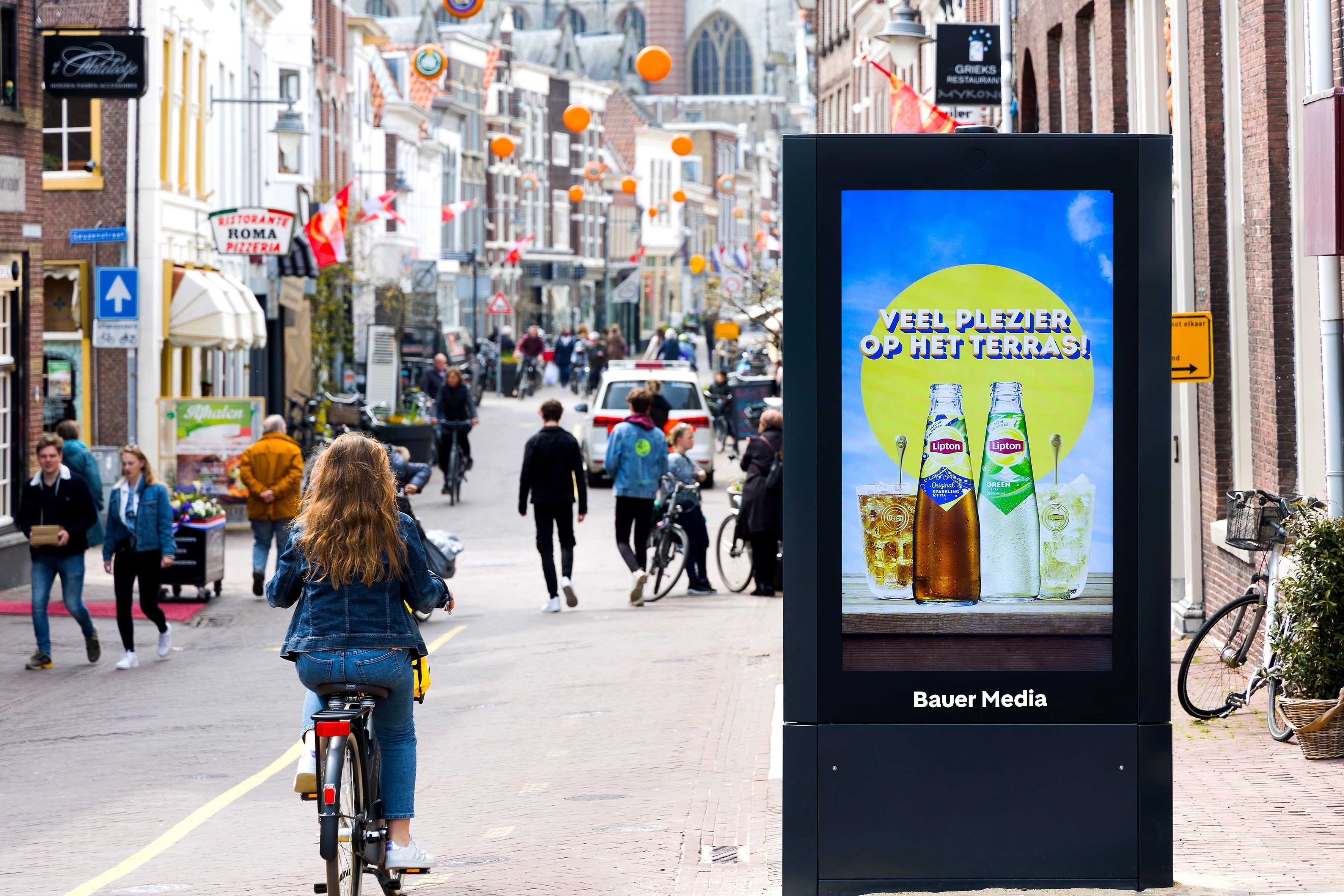 Digitaal scherm met Ice Tea reclame, vrouw die voorbij fietst en voetgangers op achtergrond