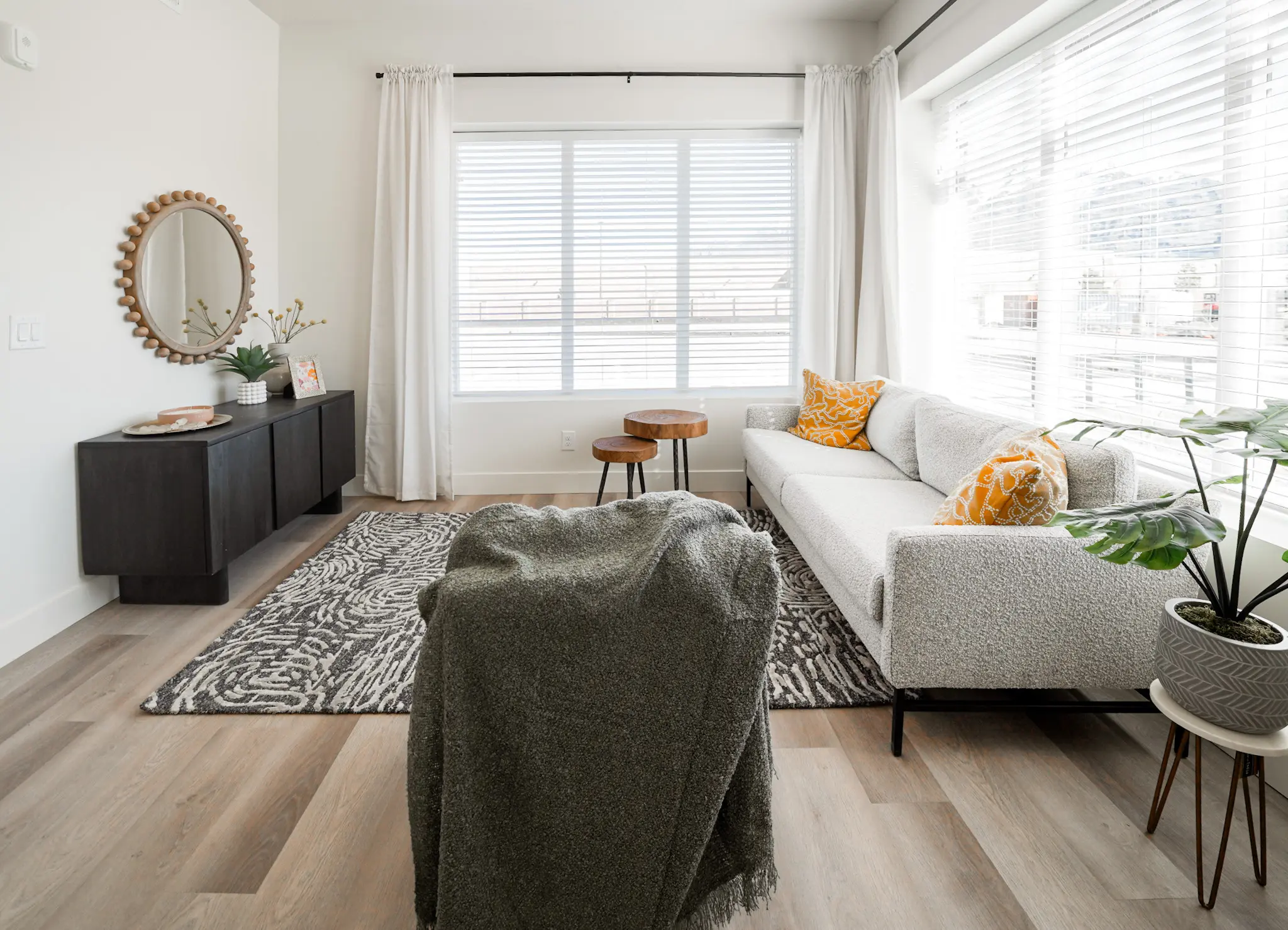 An apartment living room with stools, a gray couch with orange pillows, a chair with a blanket on it and a black wooden cupboard.