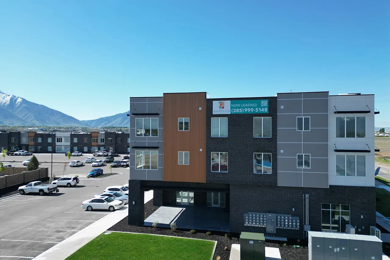 Exterior view of The Gallery Apartments in Springville, Utah, showing modern architecture, parking, and mountain scenery with a now-leasing banner.