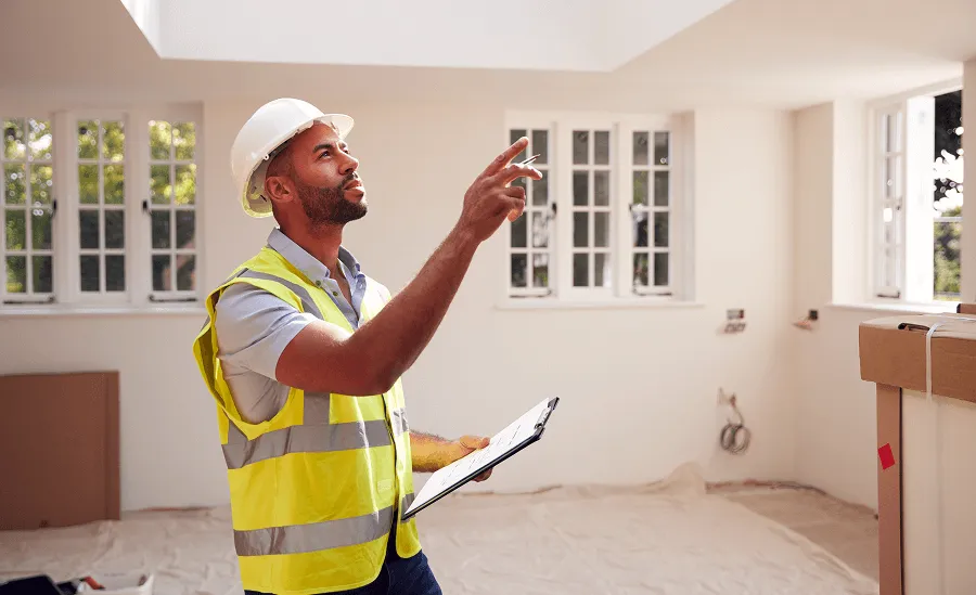 Construction worker wearing a white hard hat and yellow safety vest holding a clipboard and pointing inside a bright room under renovation.