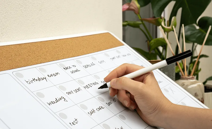 Hand holding a black marker writing 'MEETING' on a whiteboard monthly planner with events like birthday, exam, and gym.