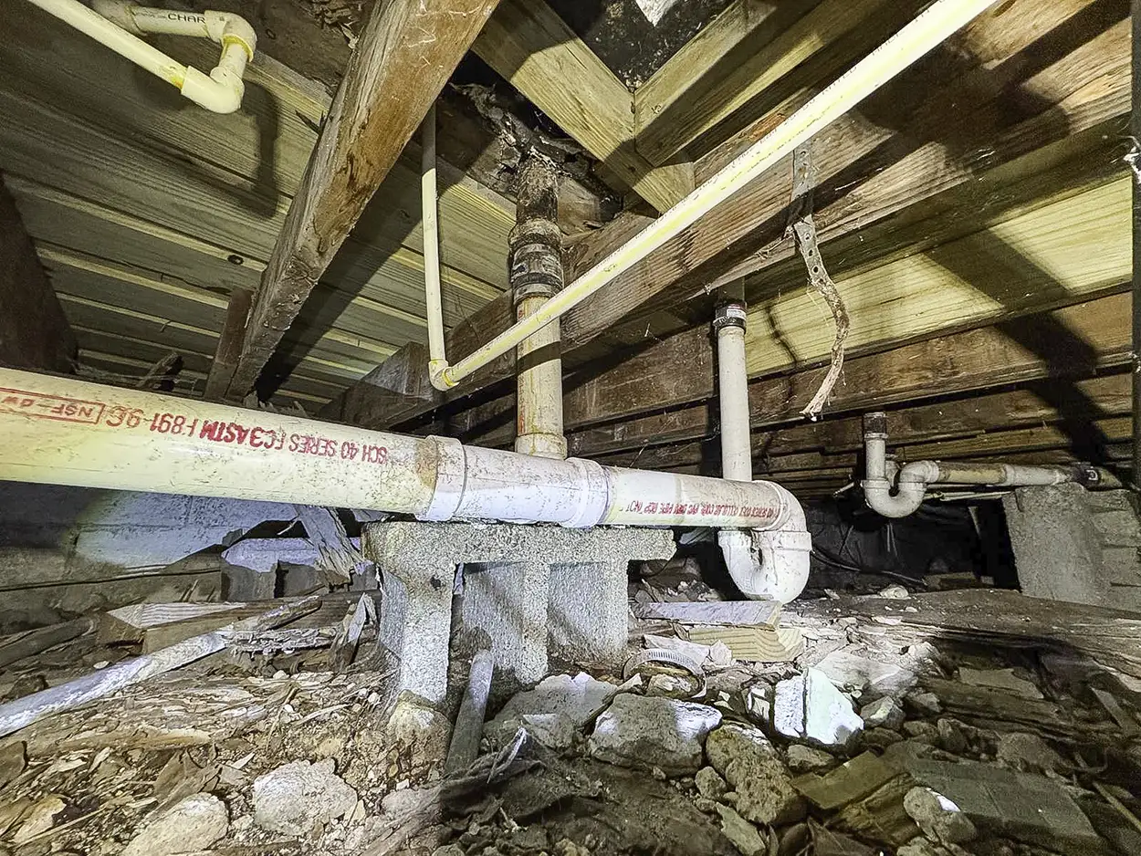 View of plumbing pipes and wooden beams in a cluttered crawl space with dirt and debris on the ground.