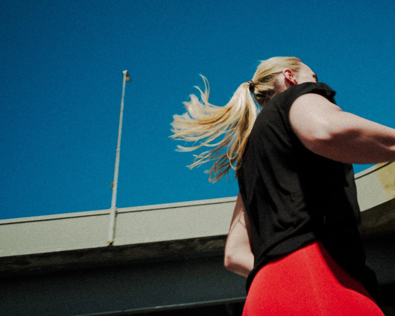 Low-angle view of a woman with blonde hair in a ponytail wearing a black top and red bottoms against a clear blue sky and concrete structure.