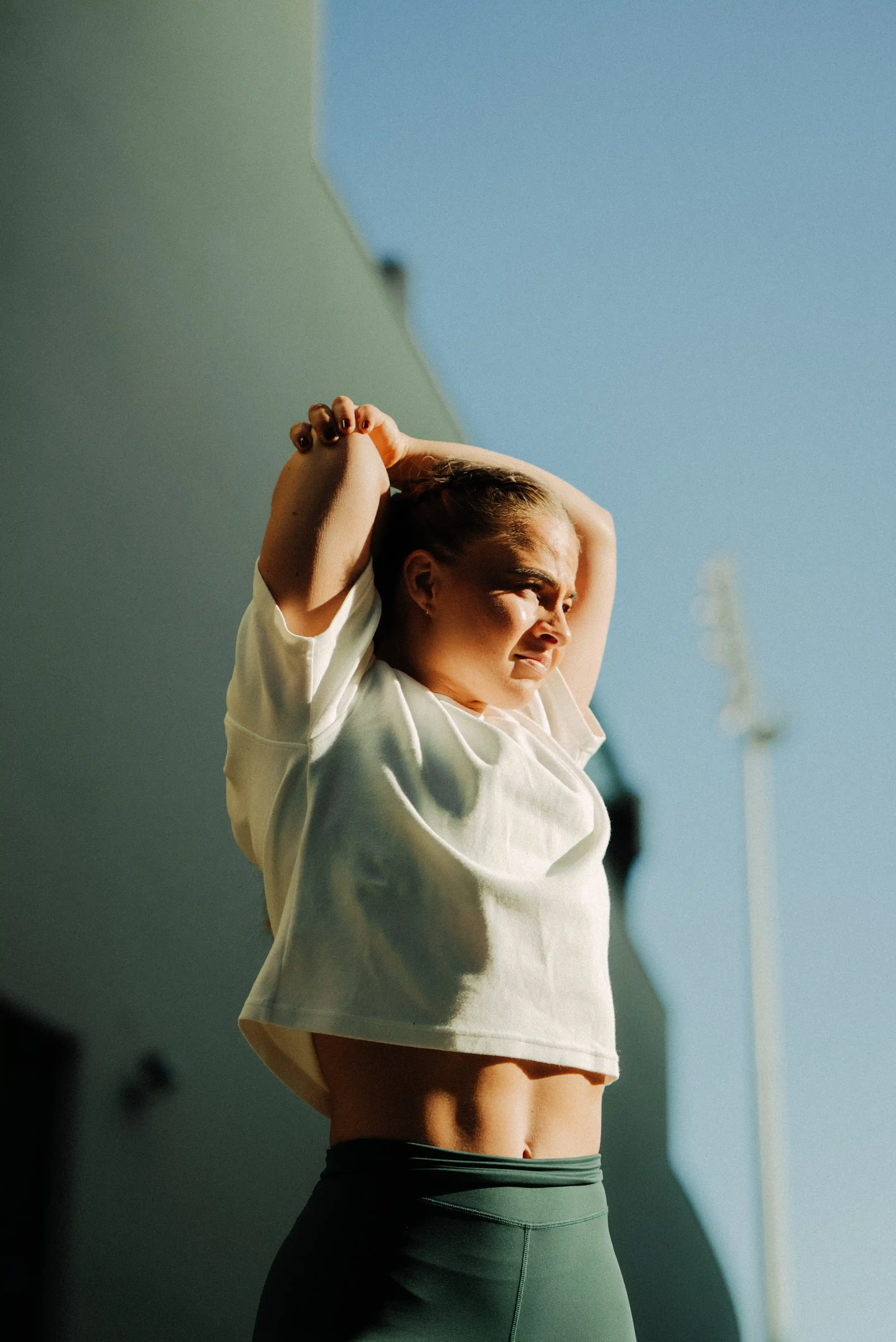 Woman in white shirt and dark green leggings stretching arms behind head outdoors in sunlight.