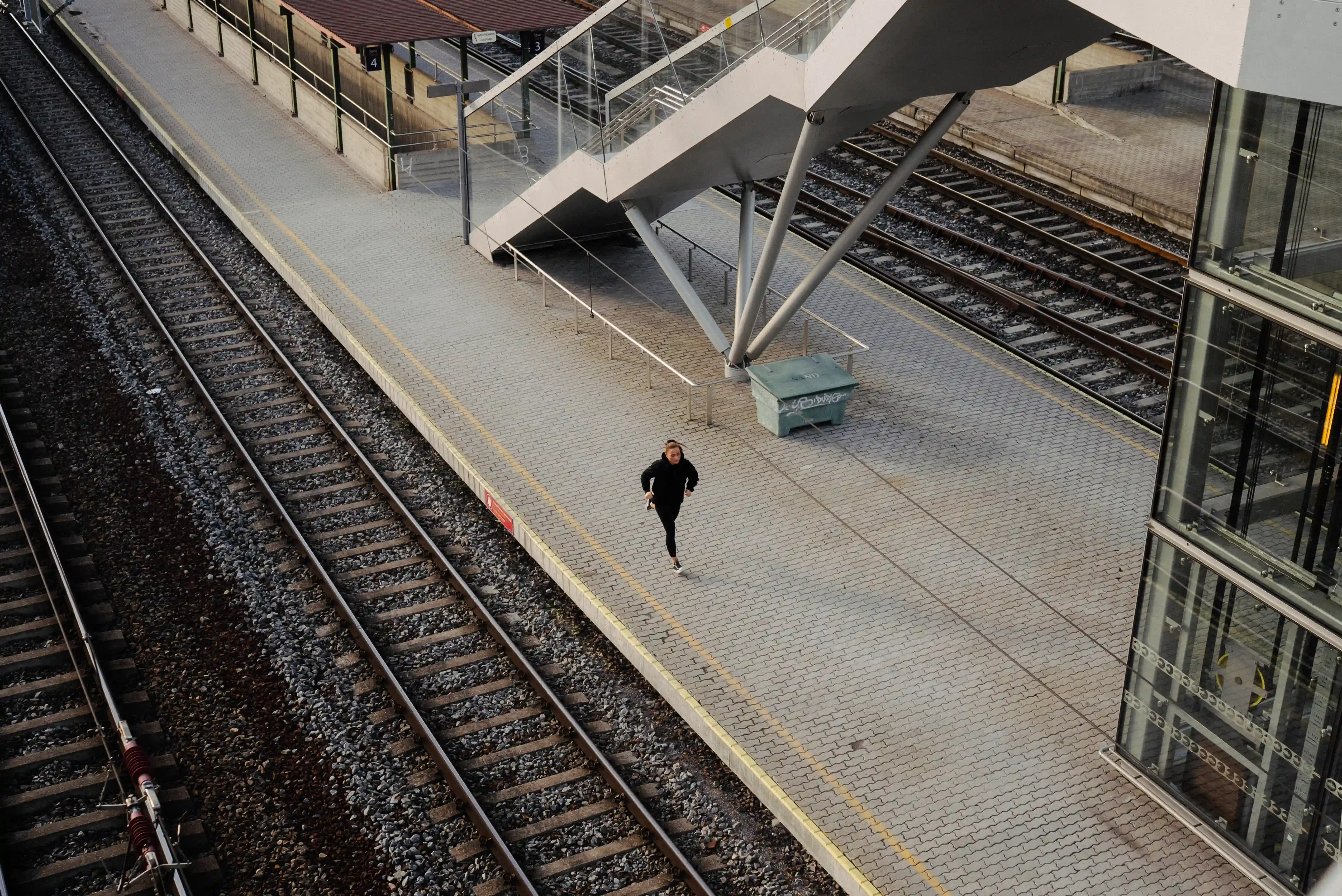 A person jogging alone on a deserted train station platform near train tracks and a modern staircase.