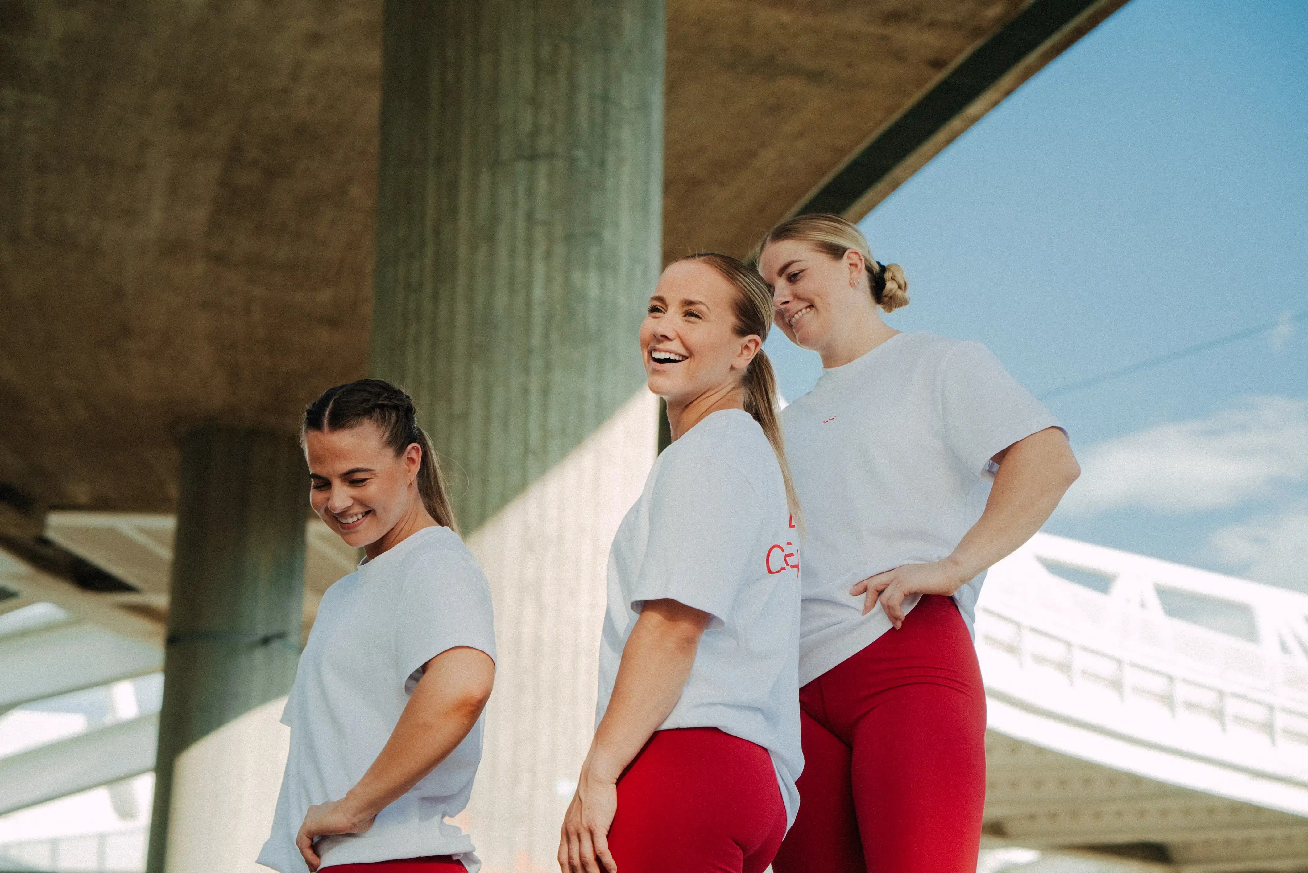 Three women in white T-shirts and red pants smiling and standing under a concrete bridge.