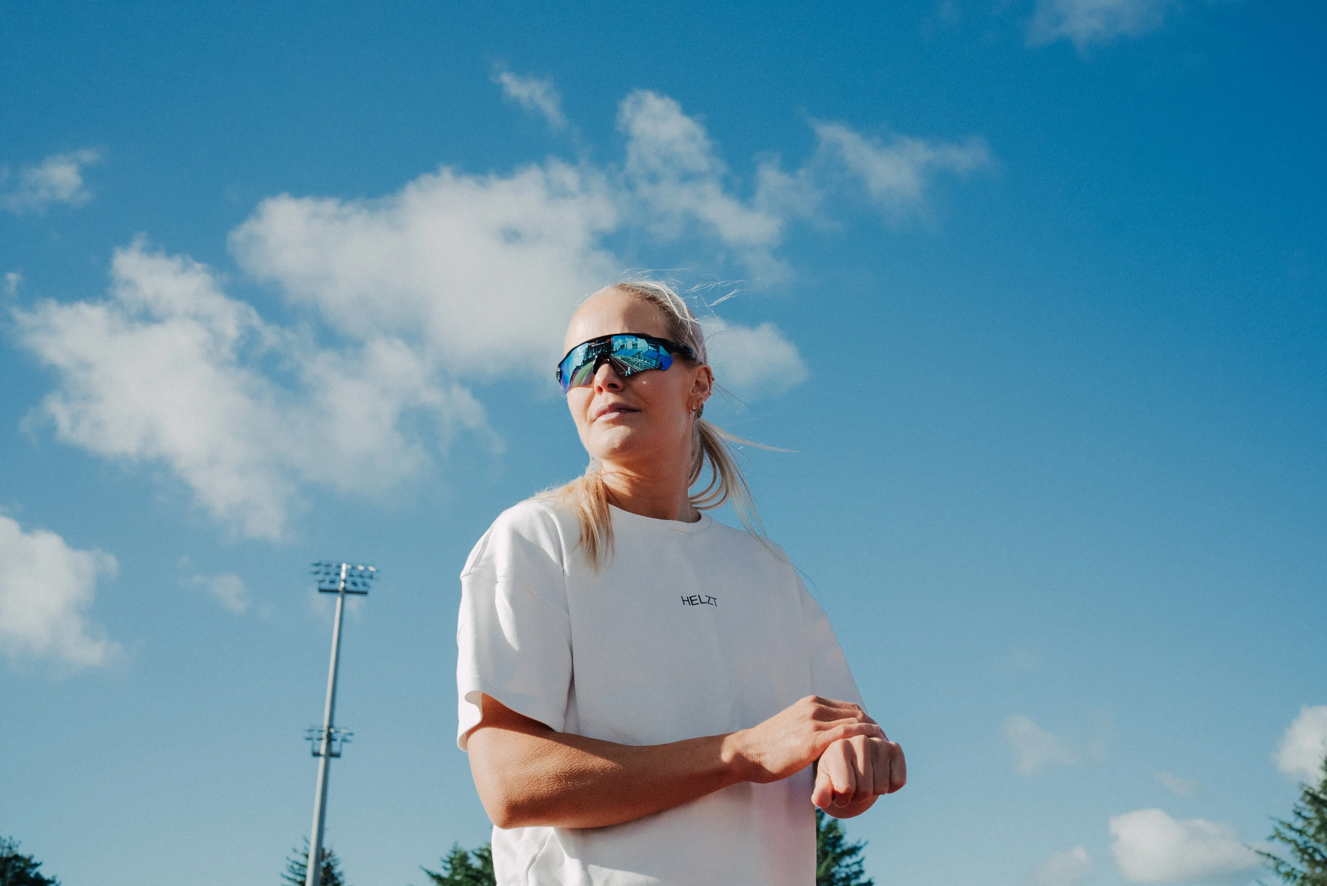 Woman in a white HELZT t-shirt and reflective sports sunglasses standing outdoors under blue sky with clouds and sports lights in the background.