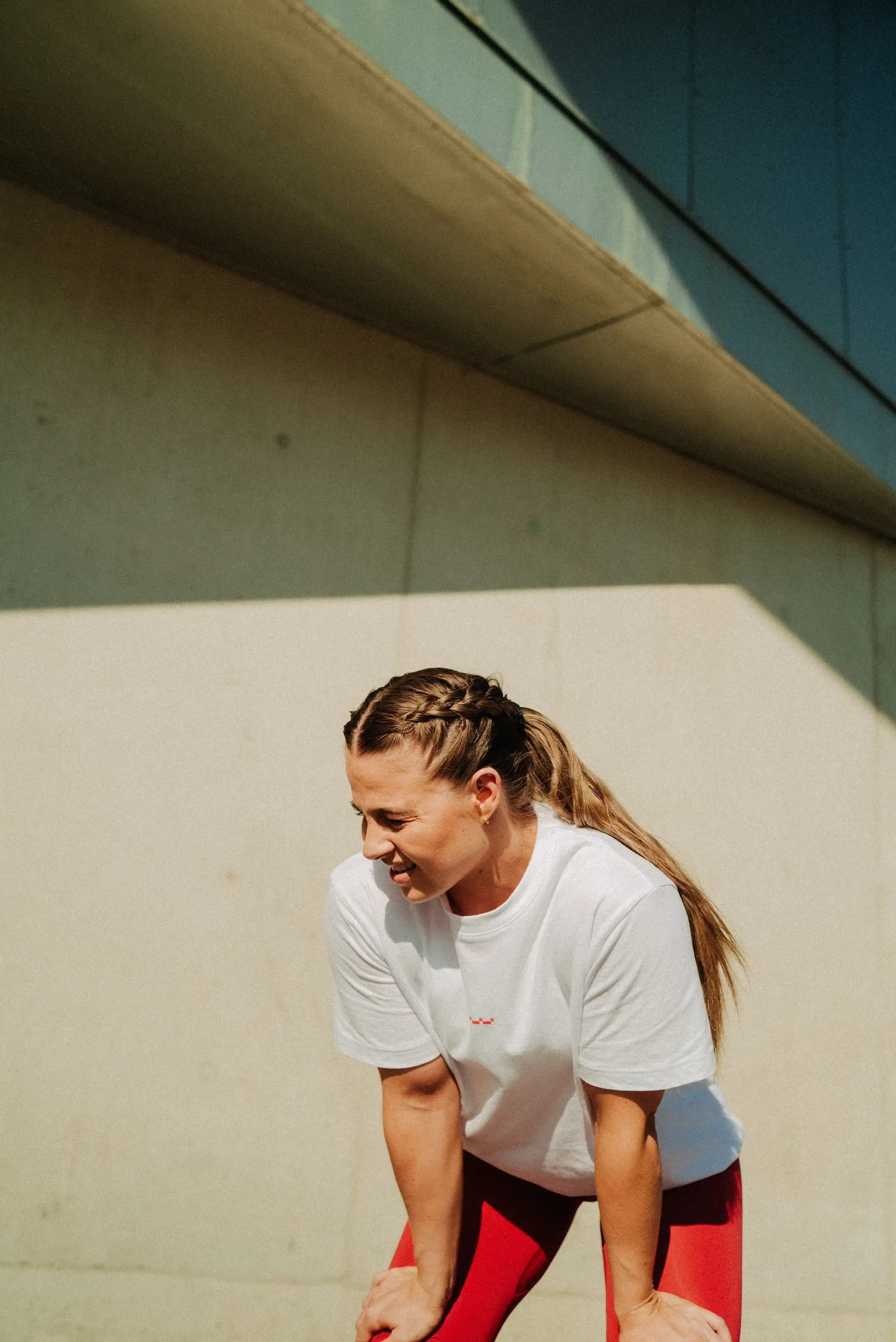 Woman with braided hair in white t-shirt and red leggings leaning forward, resting on her knees against a concrete wall.