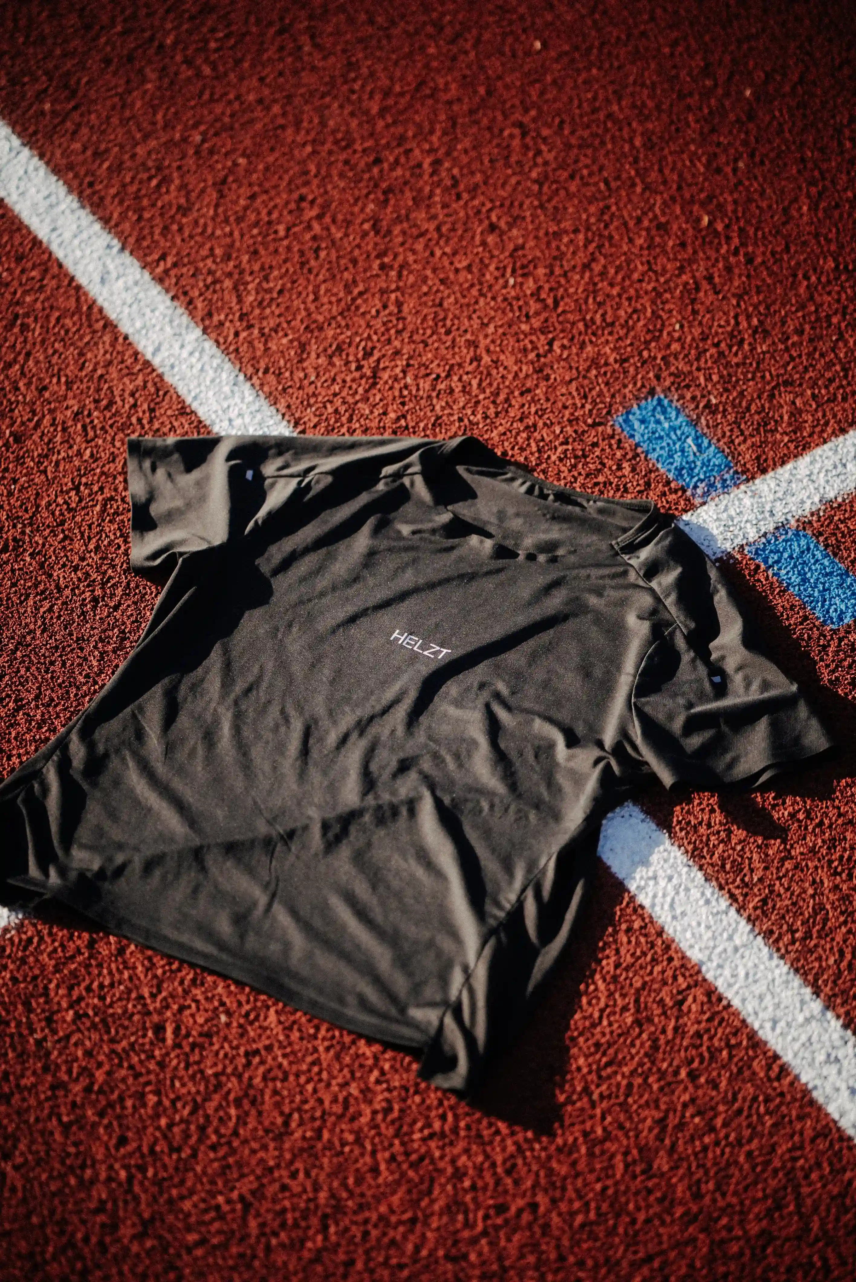 Black HELZT t-shirt laid flat on a red running track with white and blue lane markings.