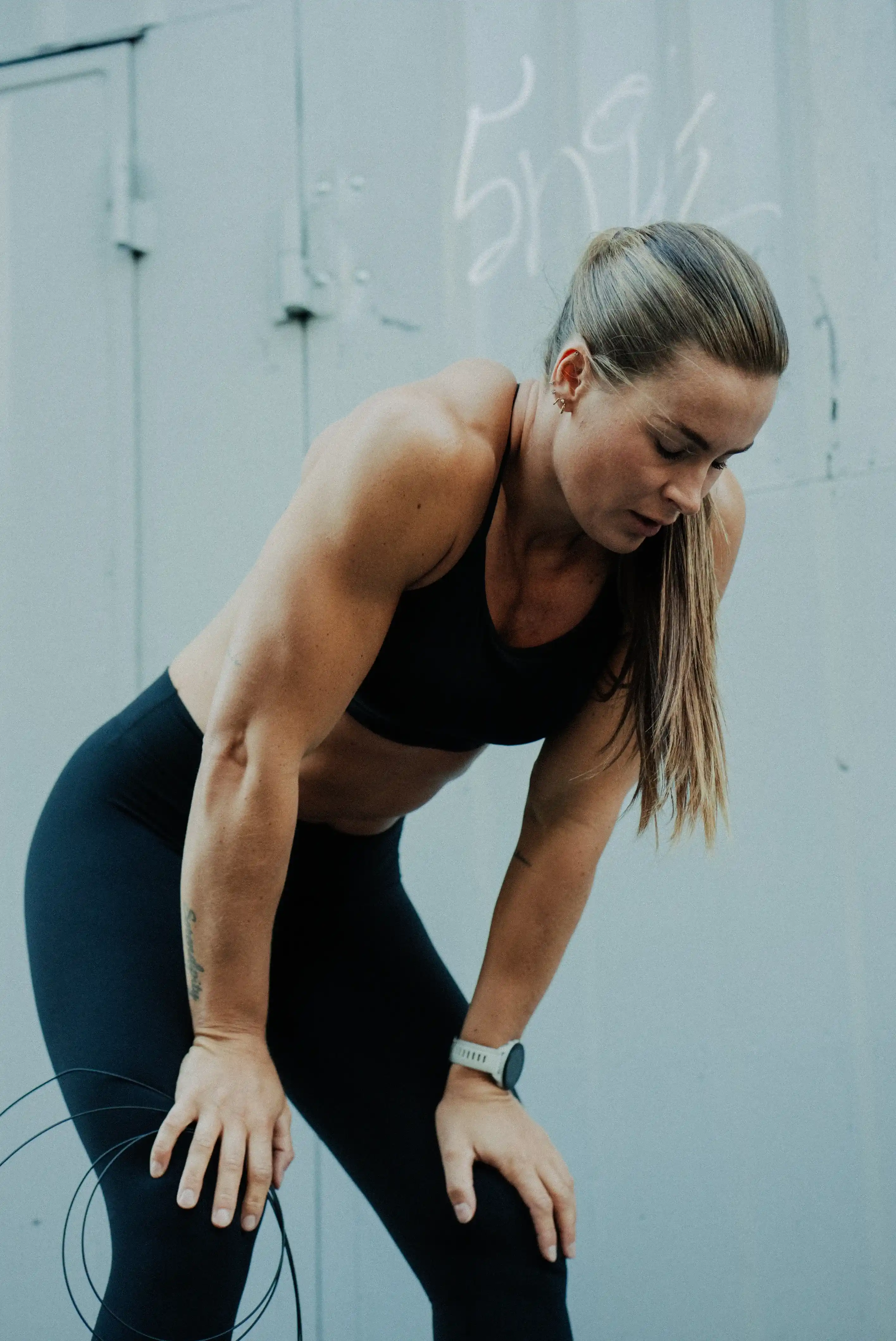 Fit woman in black workout clothes leaning forward with hands on knees, appearing tired after exercise.