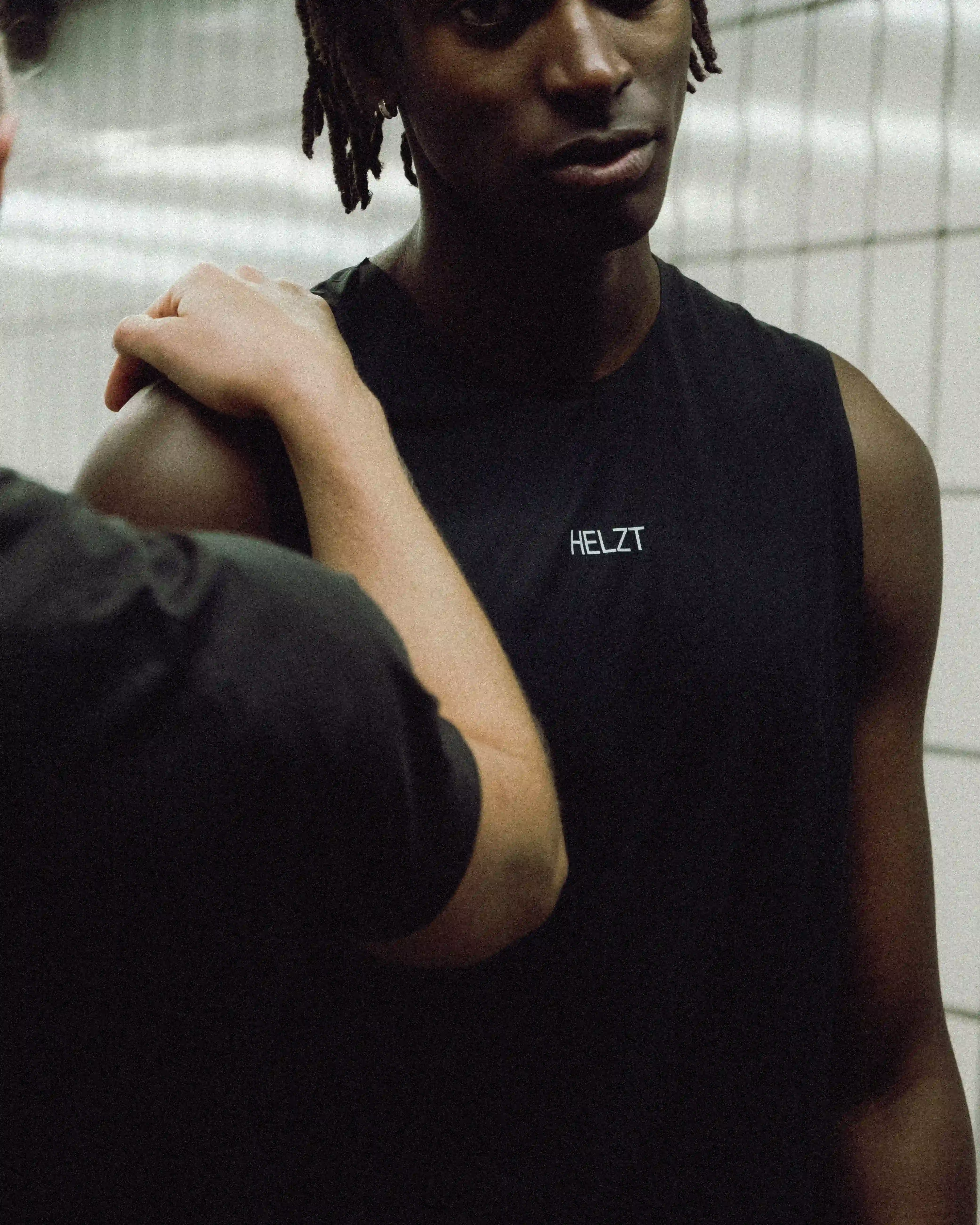 Close-up of a person wearing a sleeveless black shirt with the word 'HELZT' printed on it, while another person gently places a hand on their shoulder.