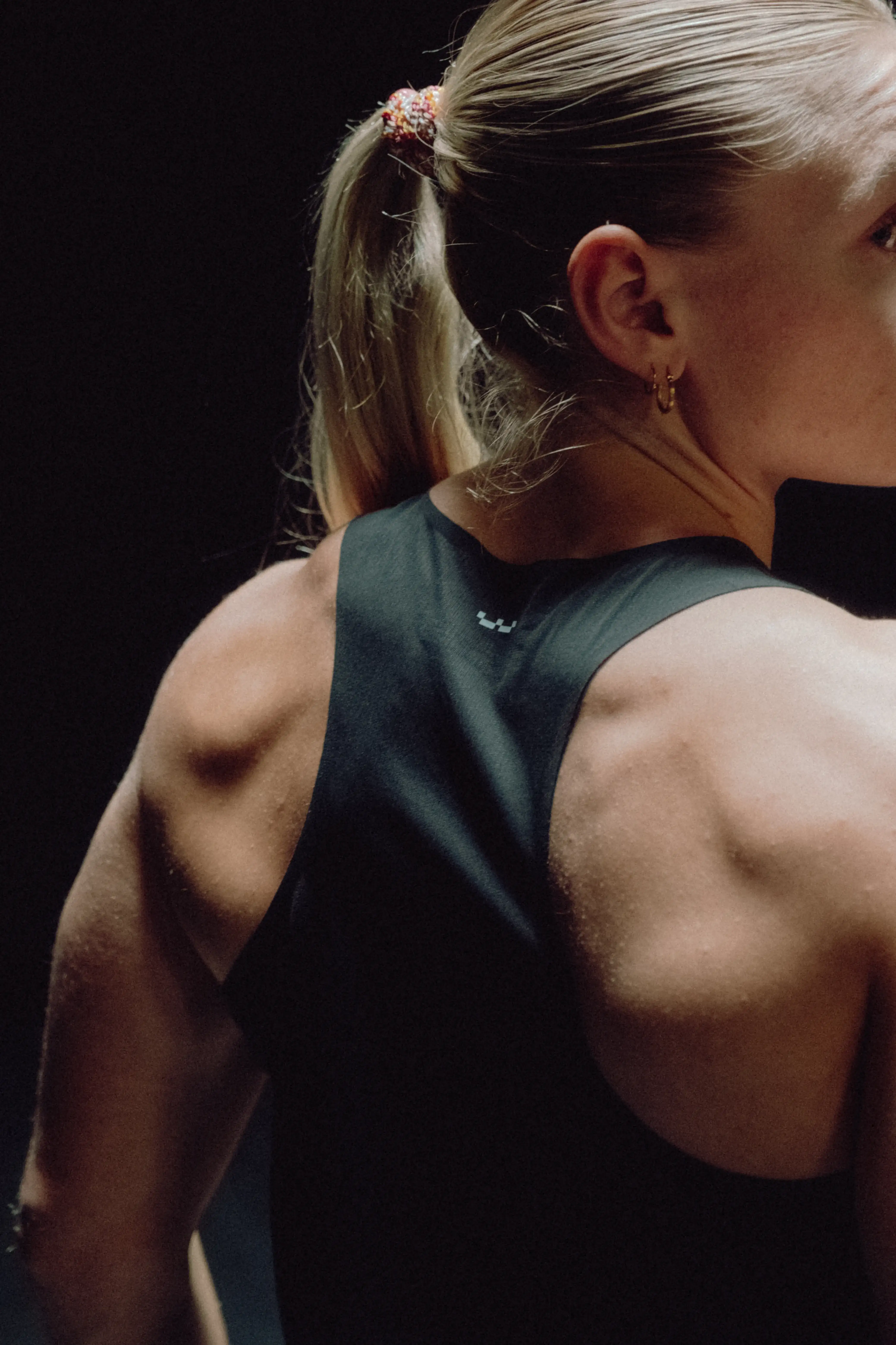 Rear view of a muscular woman with blonde hair in a ponytail wearing a black athletic tank top against a dark background.