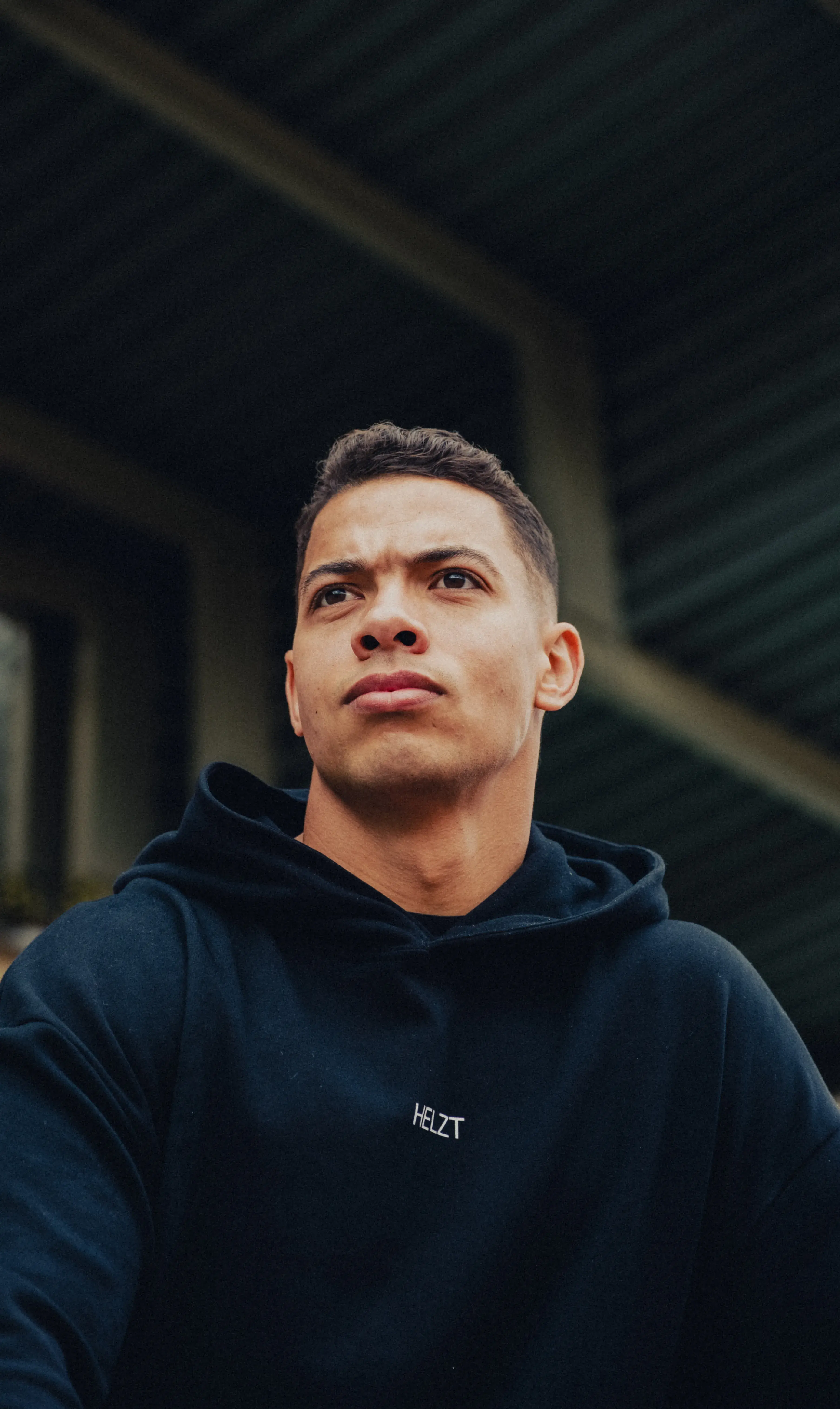Young man in a black hoodie with the word HELZT looks thoughtfully into the distance against a dark industrial background.