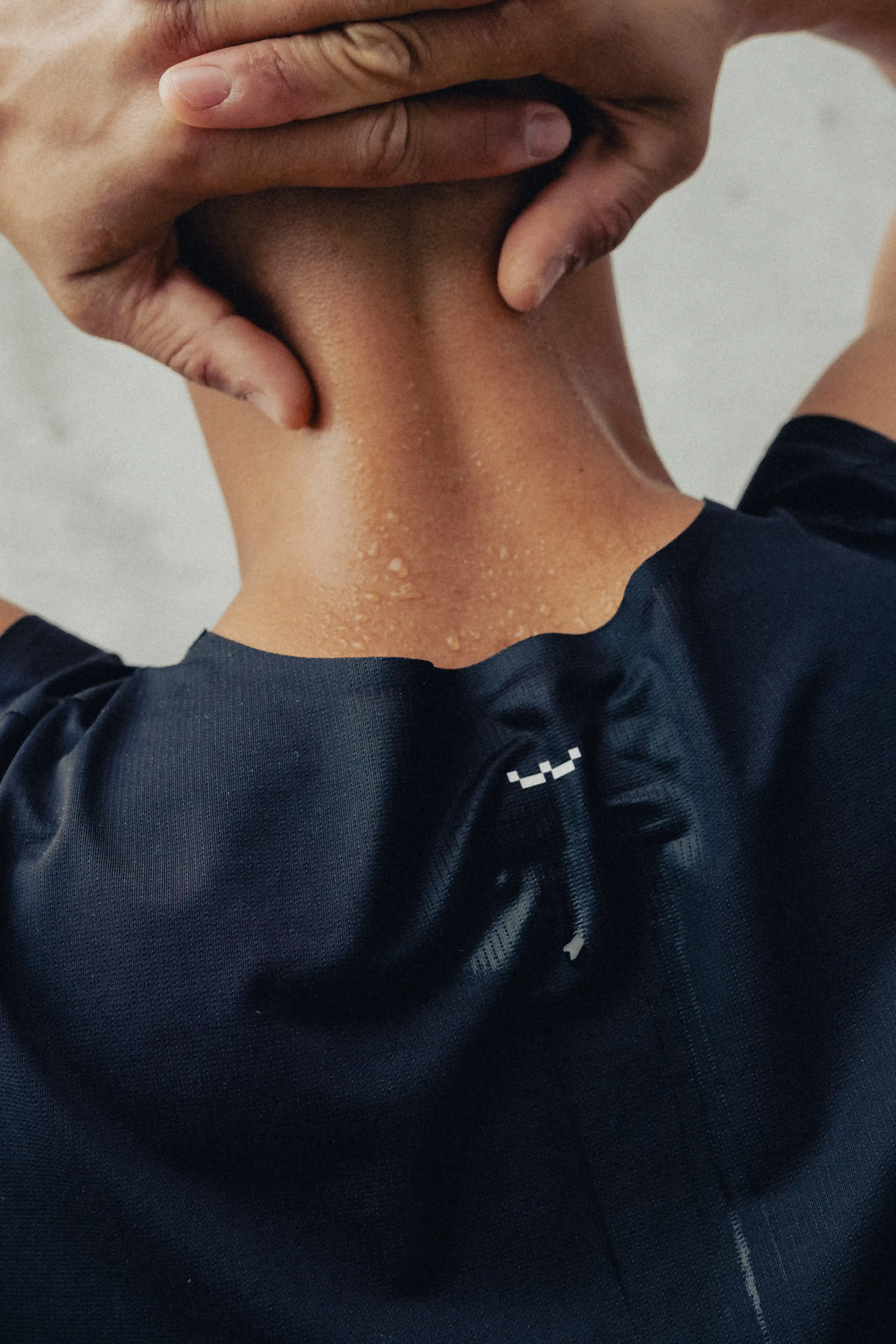 Close-up of a person’s neck and upper back with hands resting on the back of the head, wearing a dark shirt with a small logo.