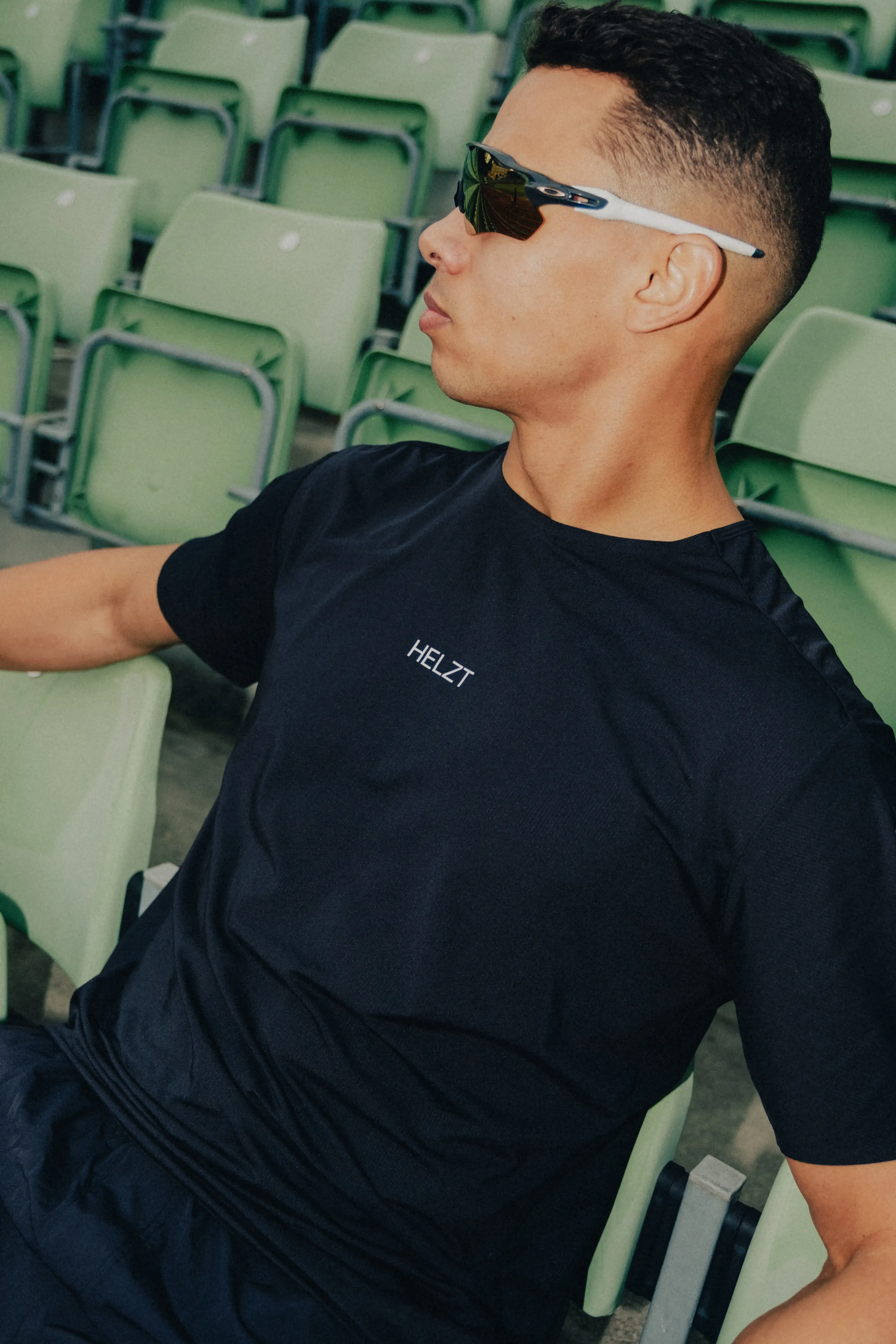 Man wearing a black HELZT t-shirt and white-framed sunglasses sitting on green stadium seats.