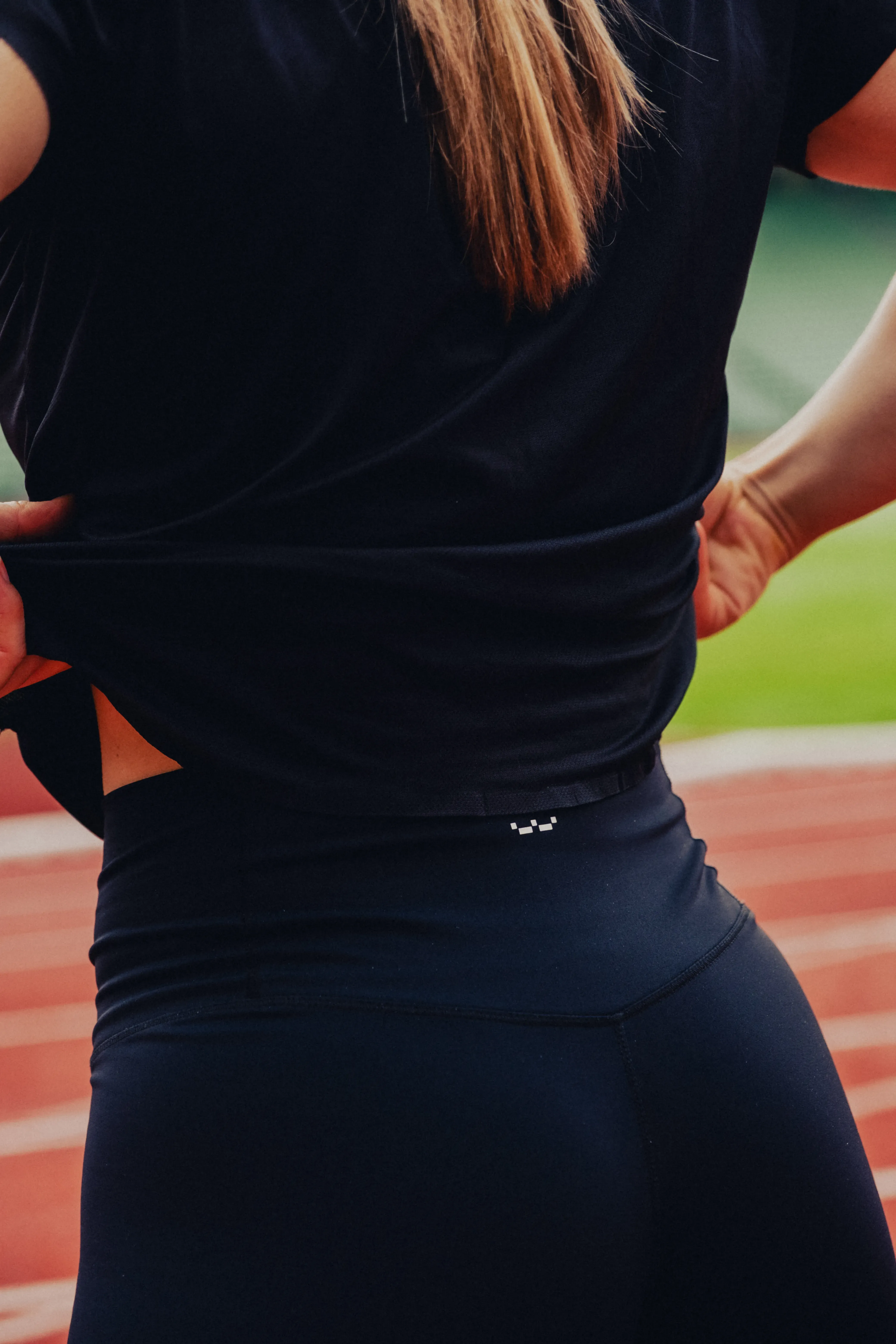 Close-up of a person lifting their black shirt at the waist, wearing black leggings, standing on a red running track.