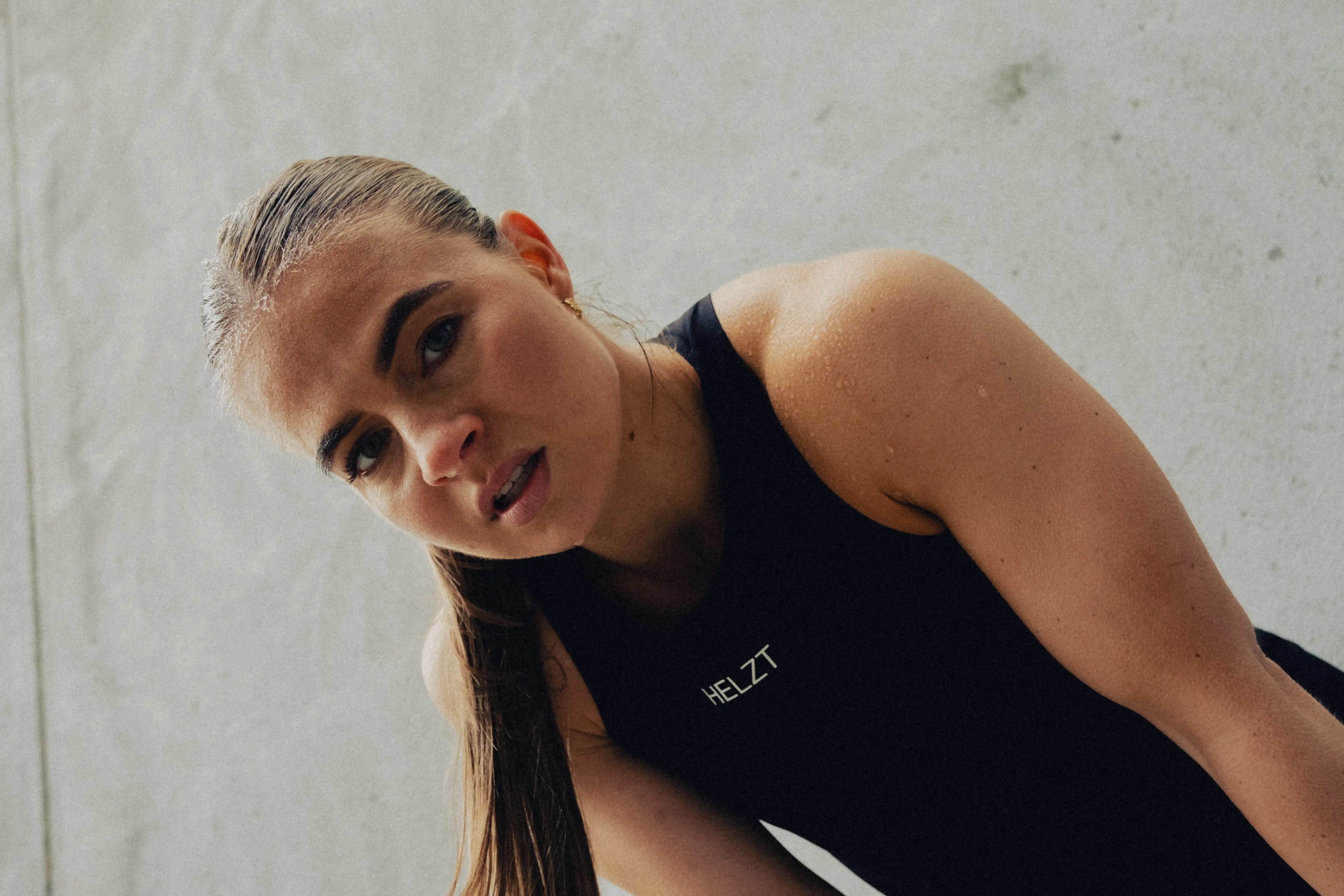 Close-up of a focused woman with wet hair in a black HELZT tank top, leaning forward against a gray background.