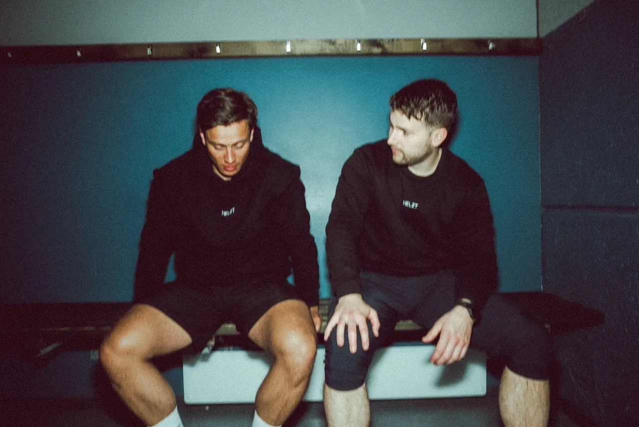 Two men sitting on a bench in a locker room, both wearing black sweatshirts.