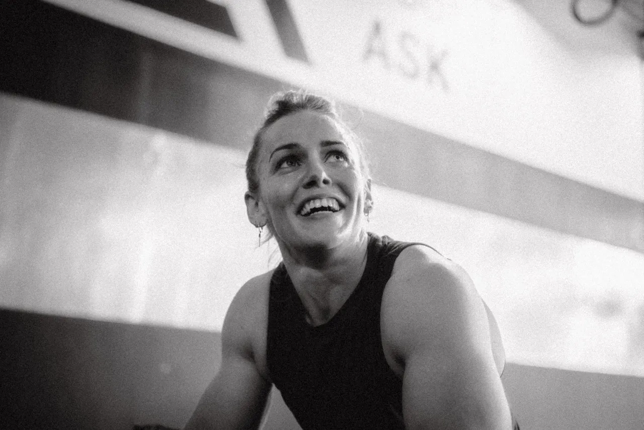 Smiling muscular woman with hair tied back, wearing a sleeveless top in a gym setting.