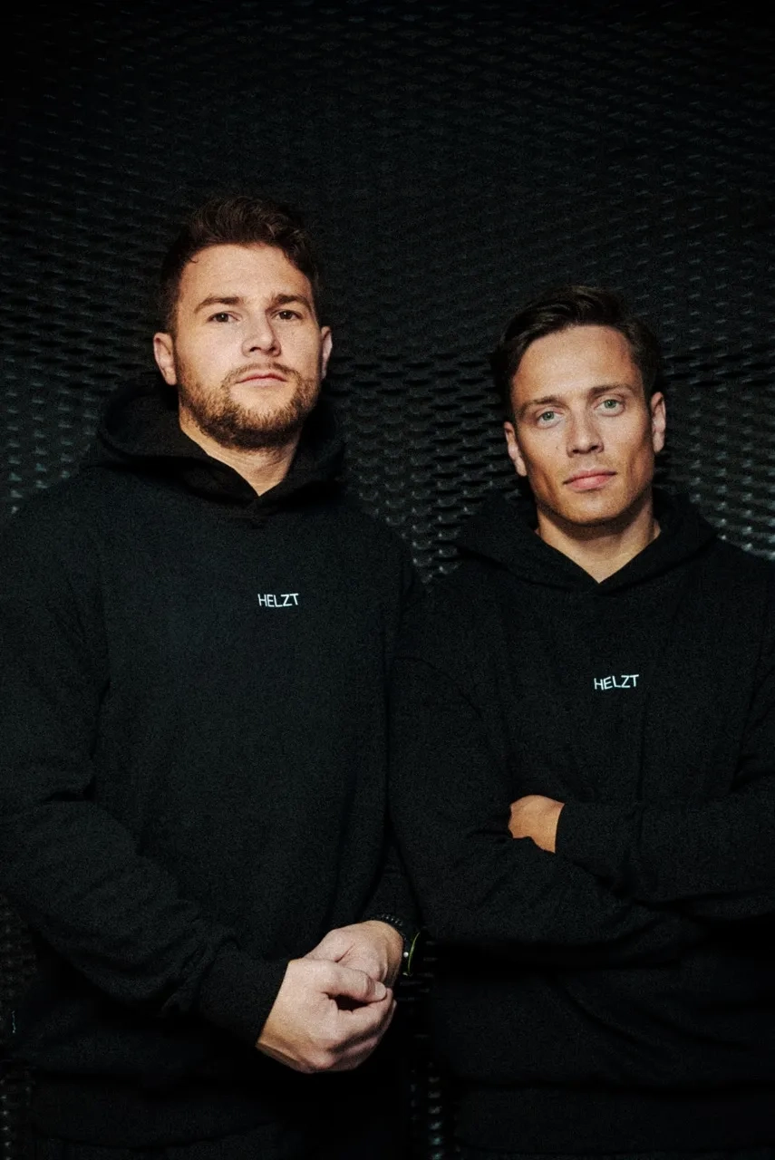 Two men wearing black hoodies with the word HELZT printed on the chest, standing against a textured black background.