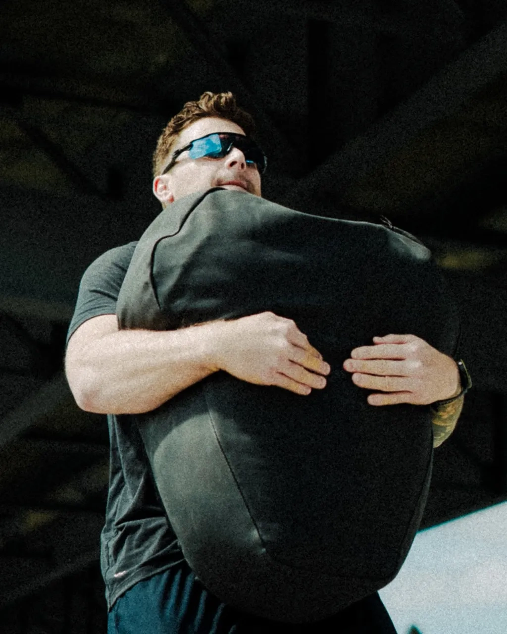 Man wearing sunglasses lifting a large black sandbag in a gym setting.