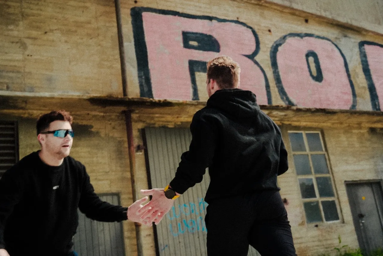 Two men dressed in black outdoors near a building with large graffiti, reaching out to shake hands.