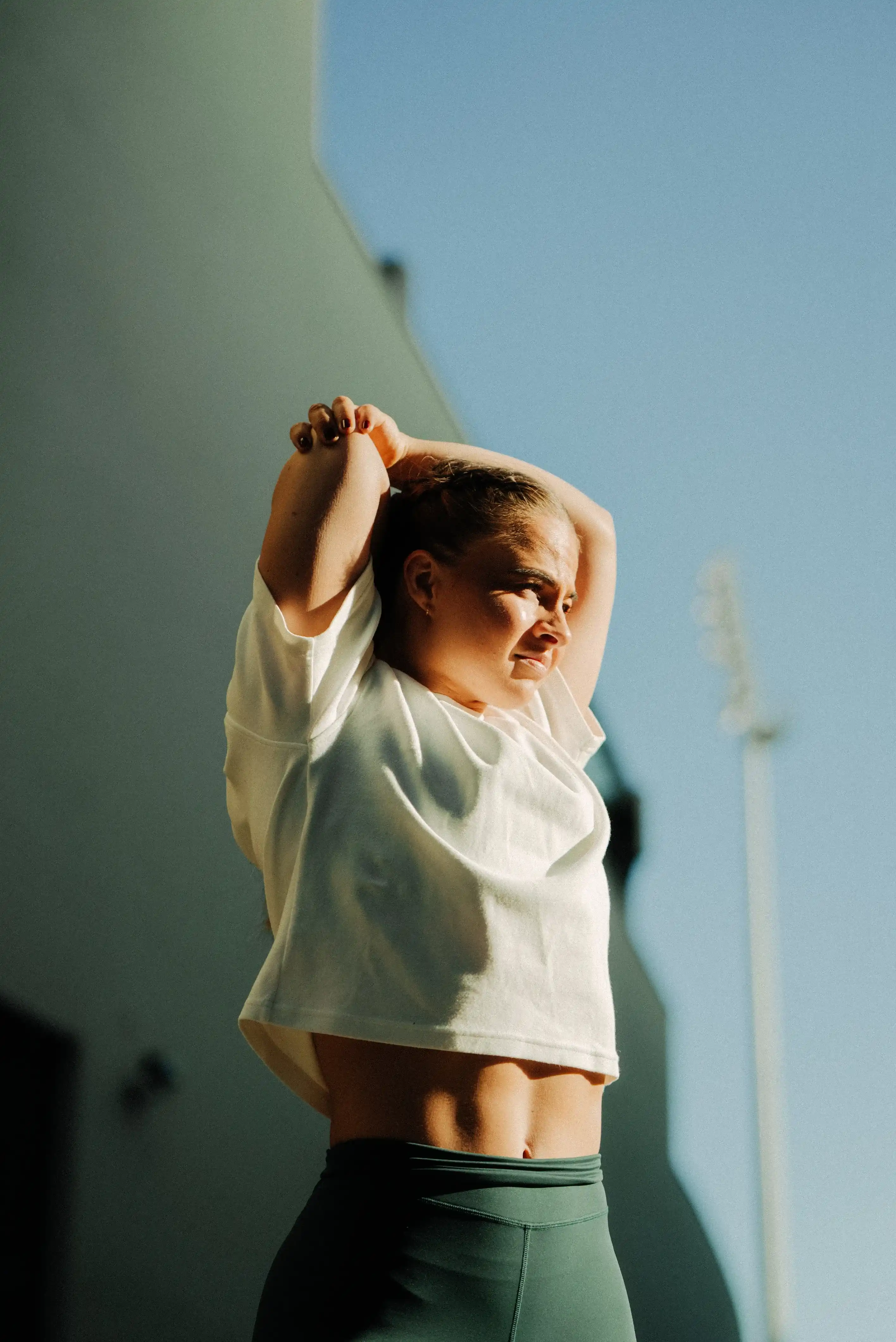 Woman in white crop top and green leggings stretching her arms overhead outdoors under clear blue sky.
