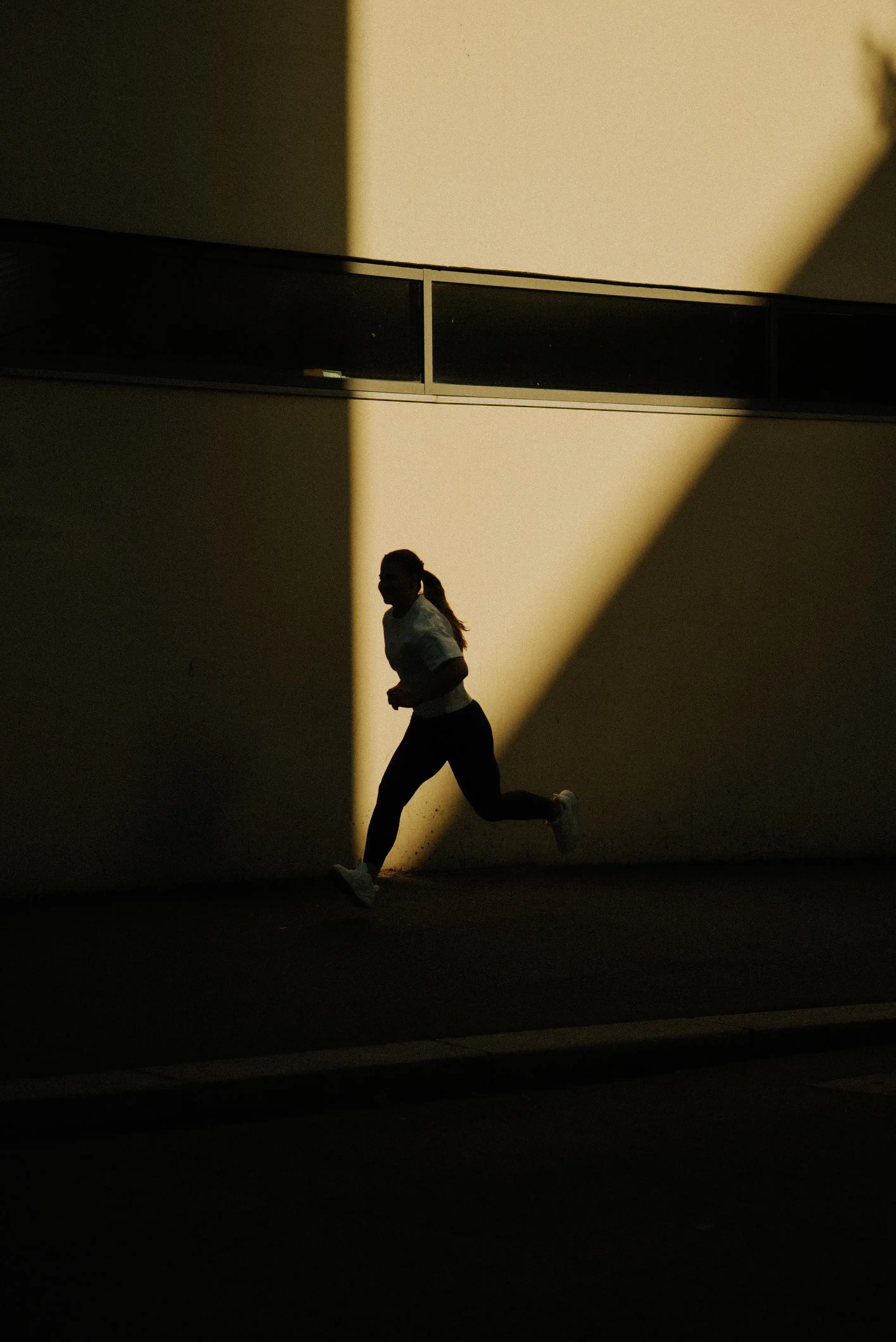 Silhouette of a person running outdoors against a wall with dramatic sunlight casting geometric shadows.
