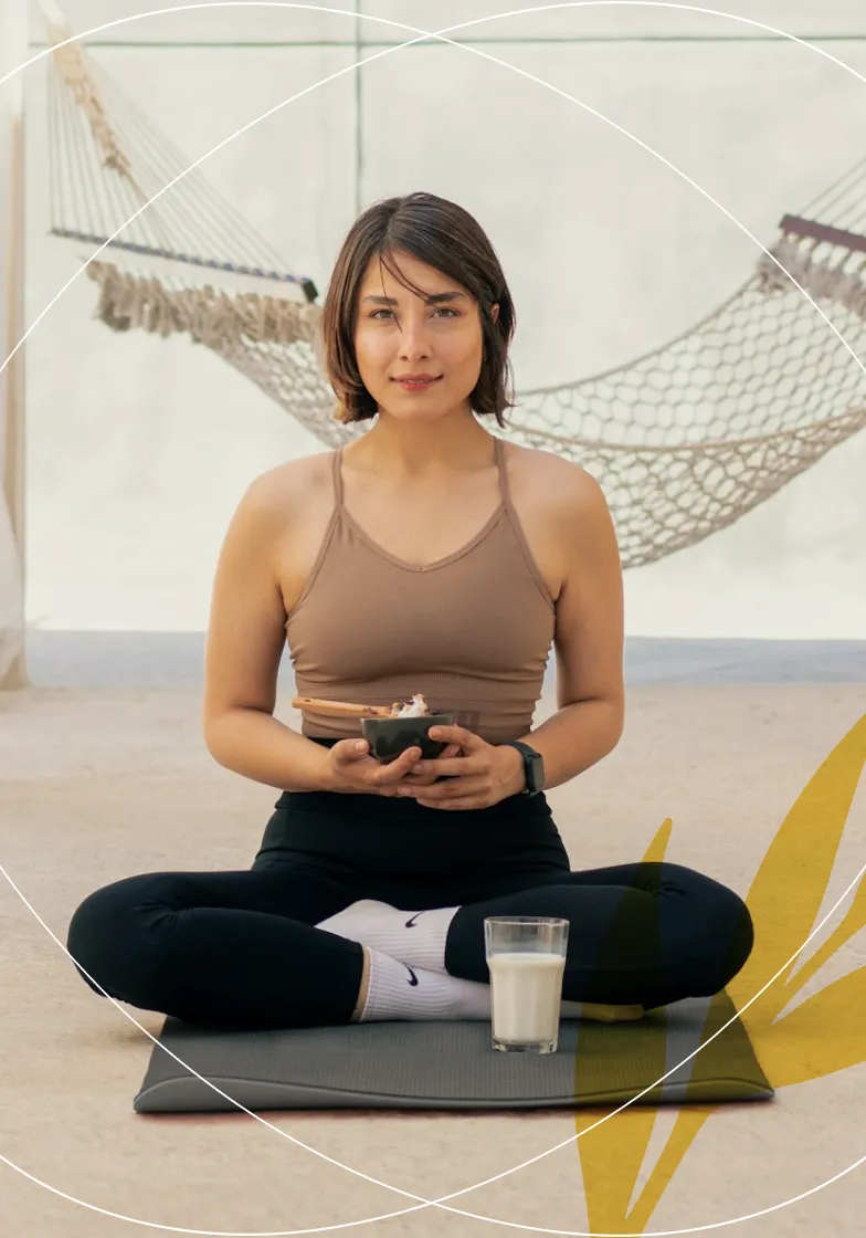 Woman seated cross-legged on a yoga mat holding a bowl with chopsticks and a glass of milk in front of her, with hammocks in the background.