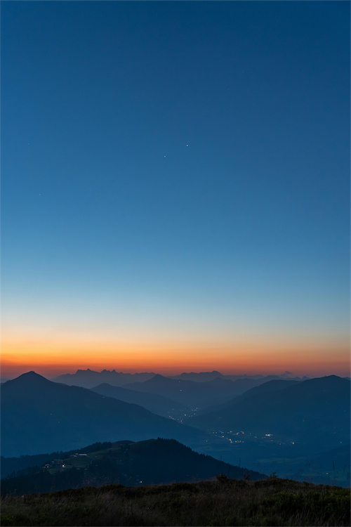 Sunset over layered mountain silhouettes with a clear sky transitioning from orange near the horizon to deep blue above.