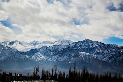 Snow-covered mountain range under a cloudy sky with silhouetted trees and buildings in the foreground.