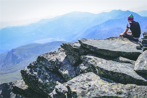 Person wearing a colorful beanie sitting on large rocky cliffs overlooking blue mountain ranges under a clear sky.