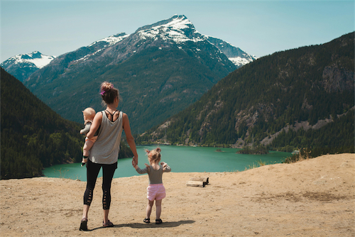 Woman holding a baby and holding hands with a toddler overlooking a lake surrounded by mountains.