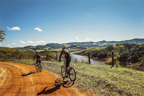 Two cyclists riding on a dirt path along a river with rolling hills under a clear blue sky.