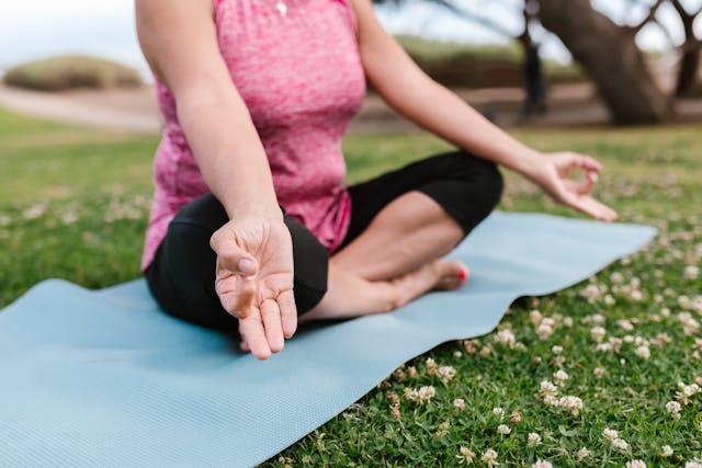 Person sitting cross-legged on a blue yoga mat on grass, meditating with hands in mudra gesture.