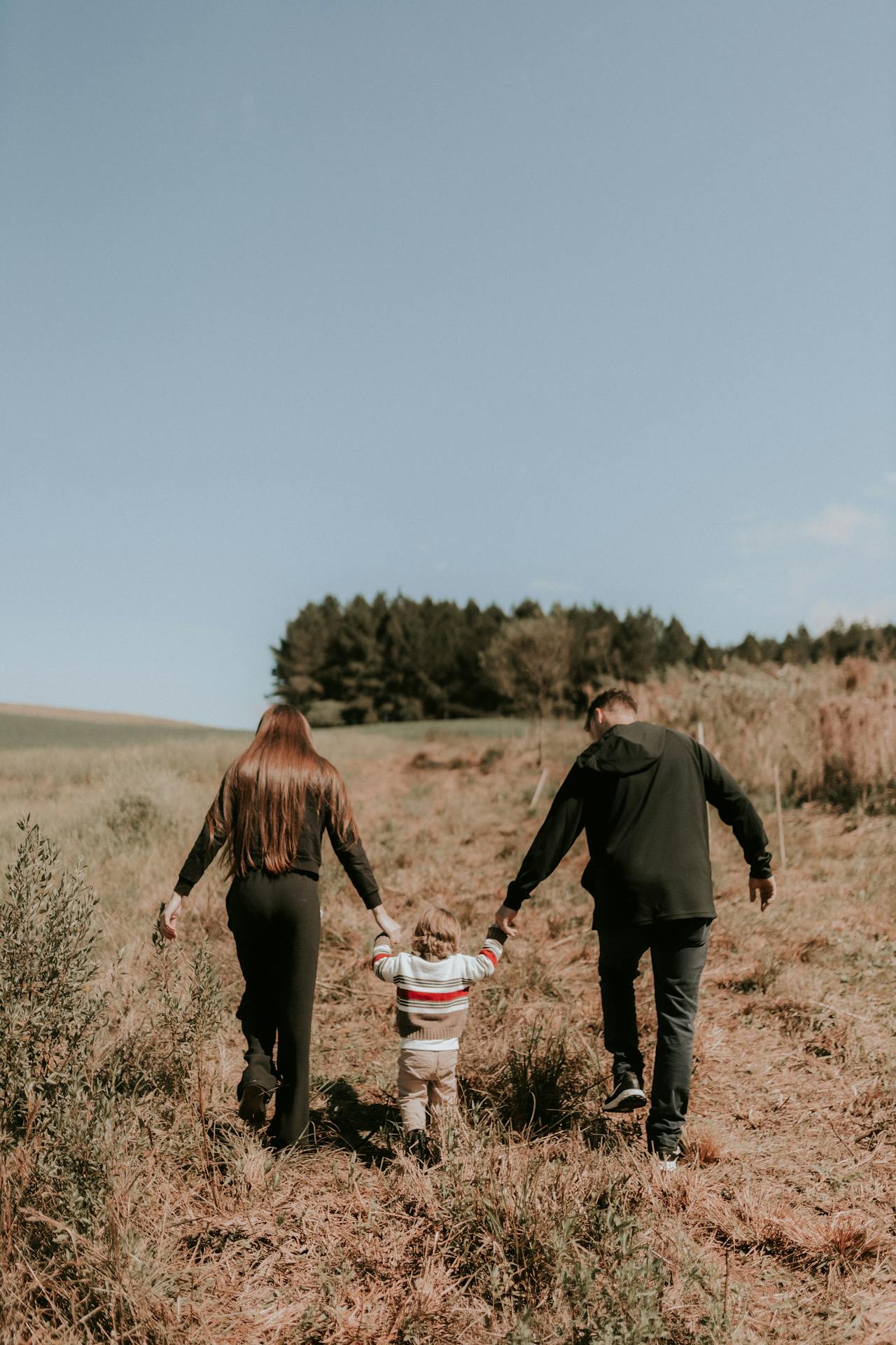 Parents holding hands with their child while walking up a grassy hill under a clear sky.