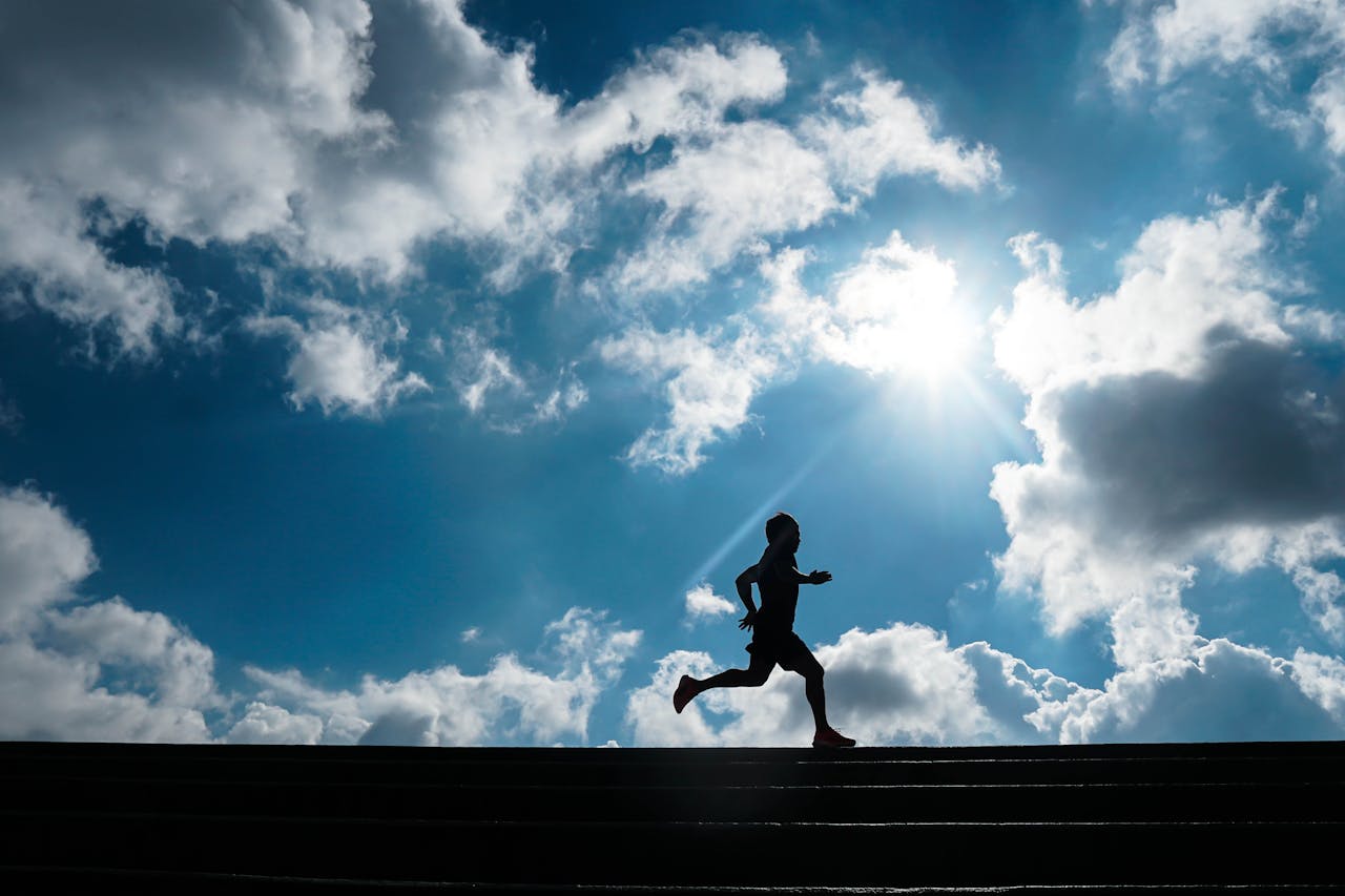 Silhouette of a person running outdoors against a bright sun and cloudy blue sky.