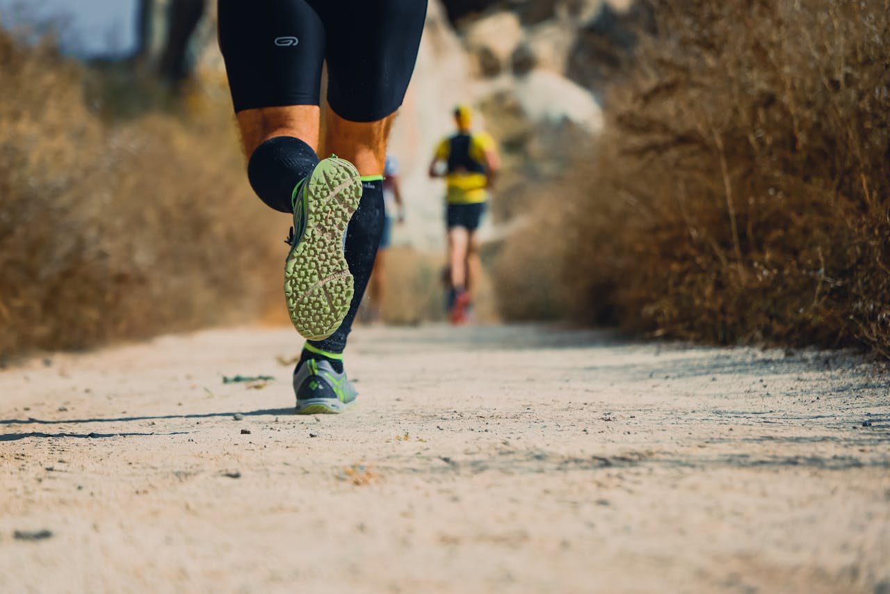 Close-up of a runner's legs and shoe soles on a dry dirt trail with other runners ahead.