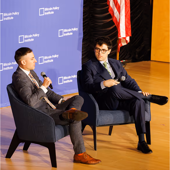 Two men in suits sitting on stage chairs holding microphones, engaged in a discussion at a Bitcoin Policy Institute event with an American flag in the background.