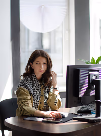 Woman with long brown hair sitting at desk using a desktop computer by a window with natural light.