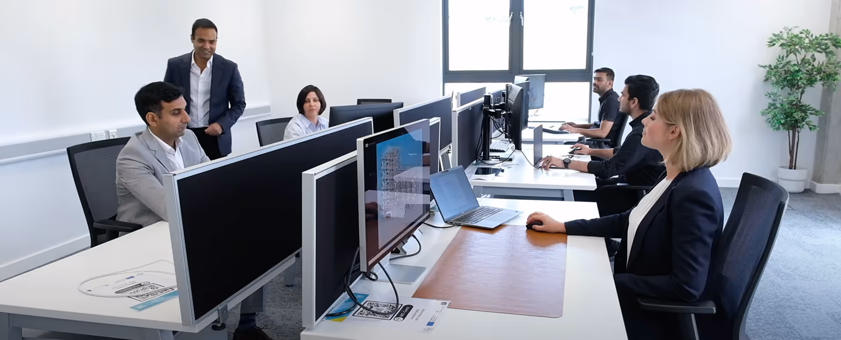 A group of six diverse professionals working at computers in a modern office with large windows and a plant in the corner.